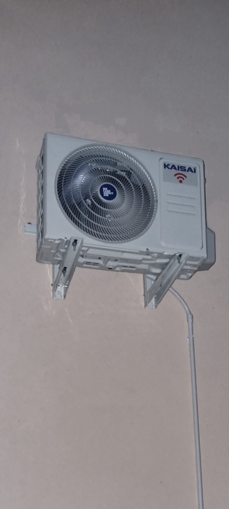 Biała jednostka wewnętrzna klimatyzatora marki Kaisai zamontowana na ścianie, widoczne metalowe wsporniki i wystająca rurka instalacyjna.