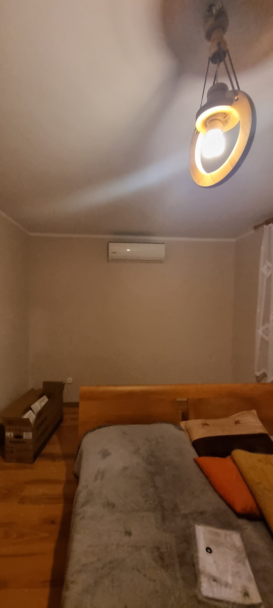 Sypialnia z zamontowanym klimatyzatorem ściennym, widoczny również drewniany zagłówek łóżka, lampa sufitowa z okrągłym kloszem i karton na podłodze.