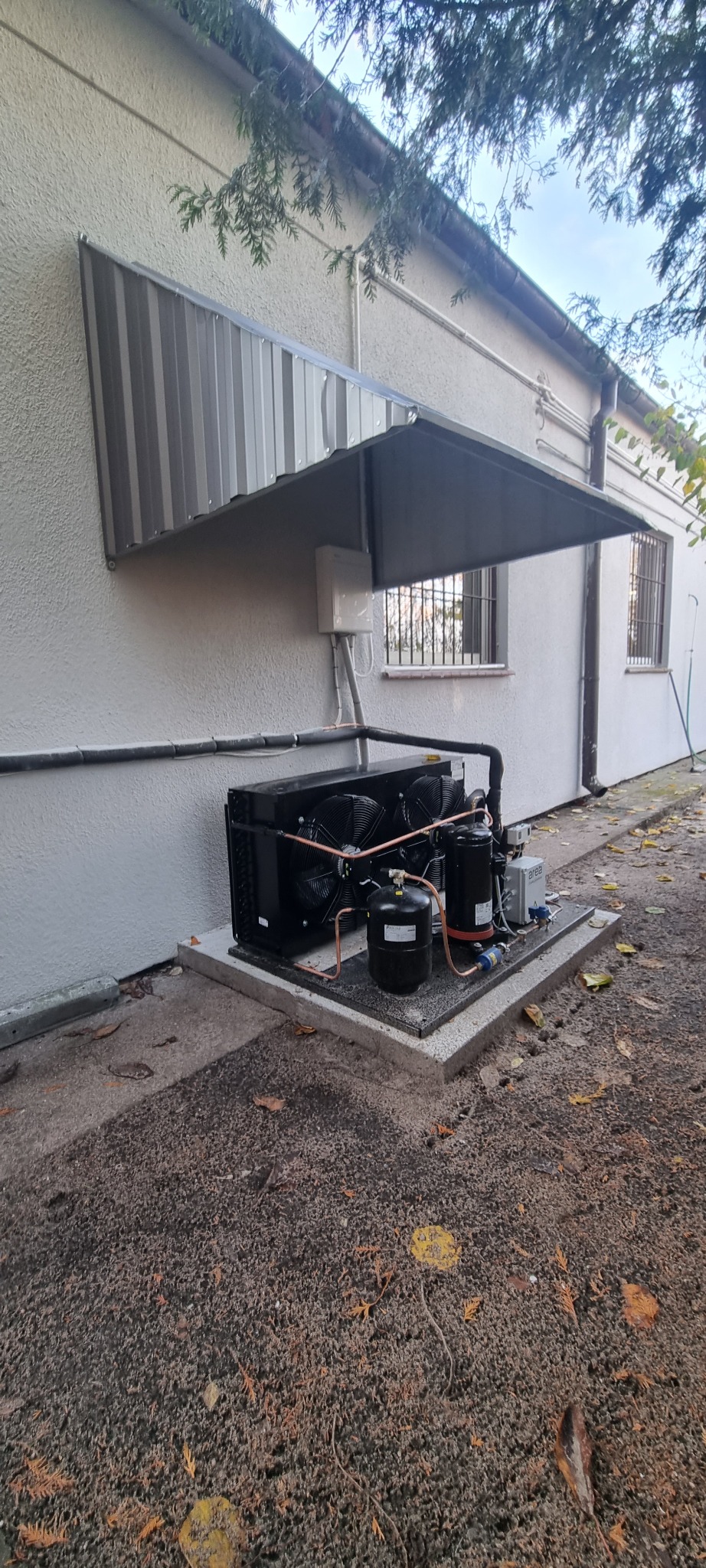 Zewnętrzna jednostka klimatyzacyjna zamontowana na betonowej podstawie przy ścianie budynku z metalowym zadaszeniem, widoczne rury miedziane i wentylatory.