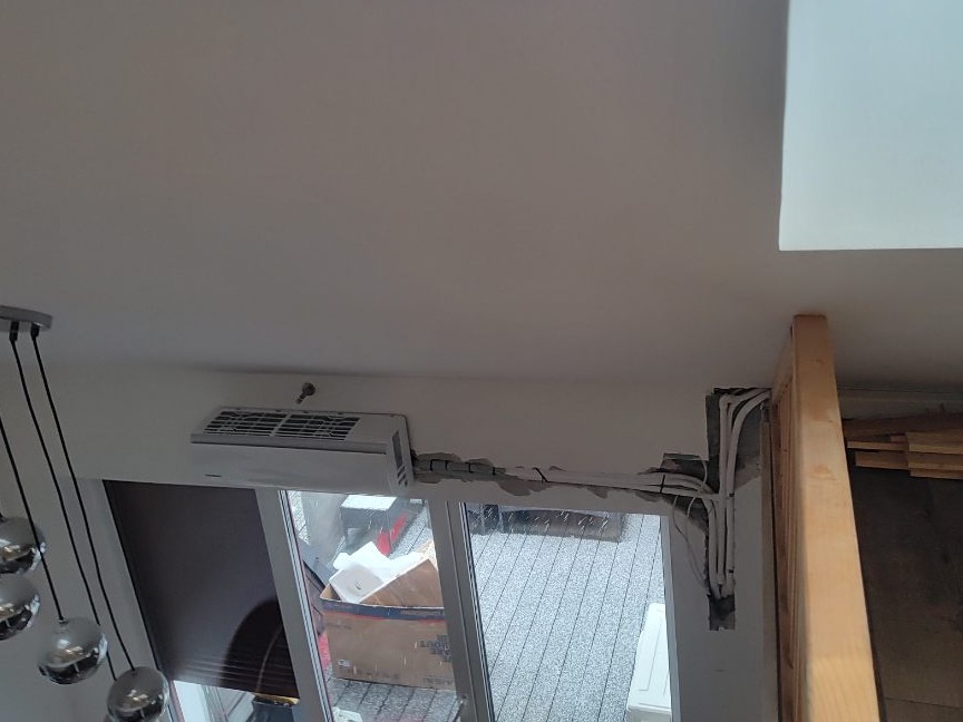 Montaż klimatyzatora w domu, widoczne przewody i otwory w ścianie, widok z góry, fragment żyrandola w lewym dolnym rogu.