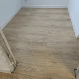 Jasne panele podłogowe imitujące drewno ułożone w pustym pokoju z białymi ścianami i listwami przypodłogowymi, widoczne surowe wykończenie ościeżnicy drzwi.