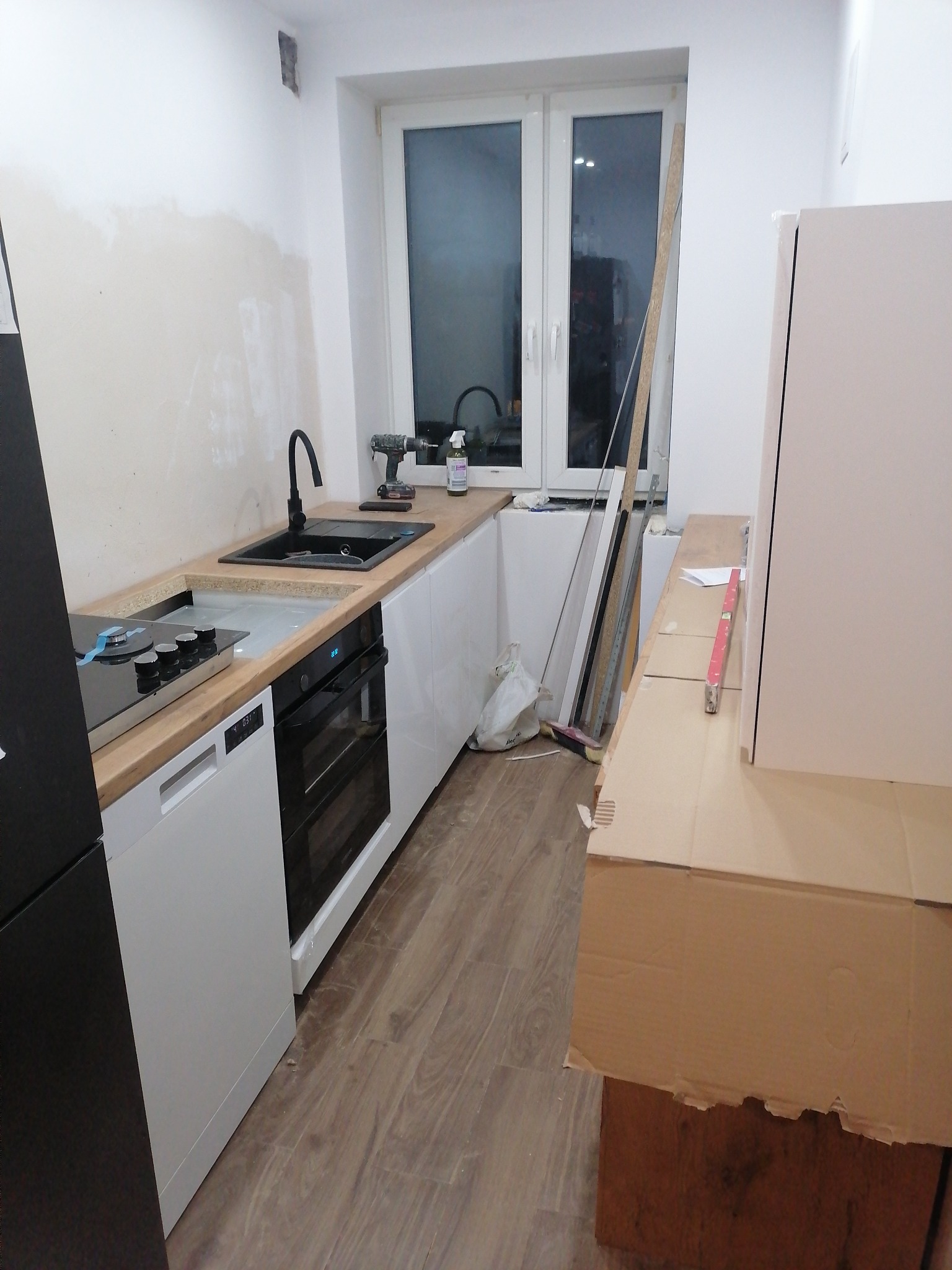 Montaż nowoczesnej kuchni: czarny zlew i bateria, płyta indukcyjna, piekarnik w zabudowie, białe fronty szafek, drewniany blat, okno w tle, widoczne narzędzia i kartony zabezpieczające.