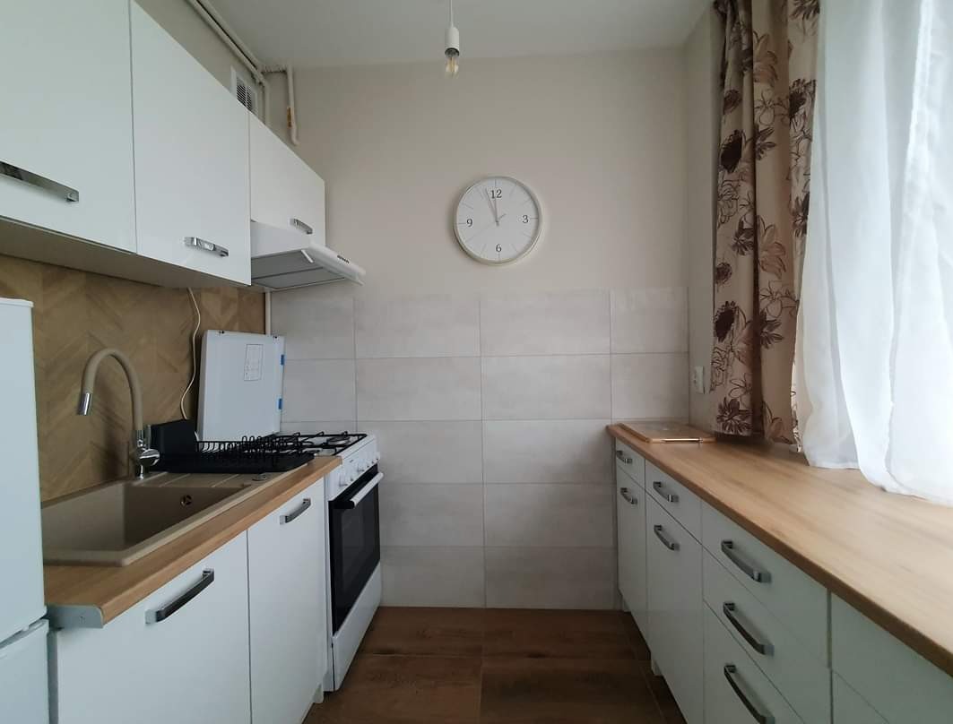 Jasna kuchnia z białymi szafkami, drewnianym blatem i beżową ścianą z zegarem. Widoczny zlew, kuchenka i okno z zasłoną w kwiaty.