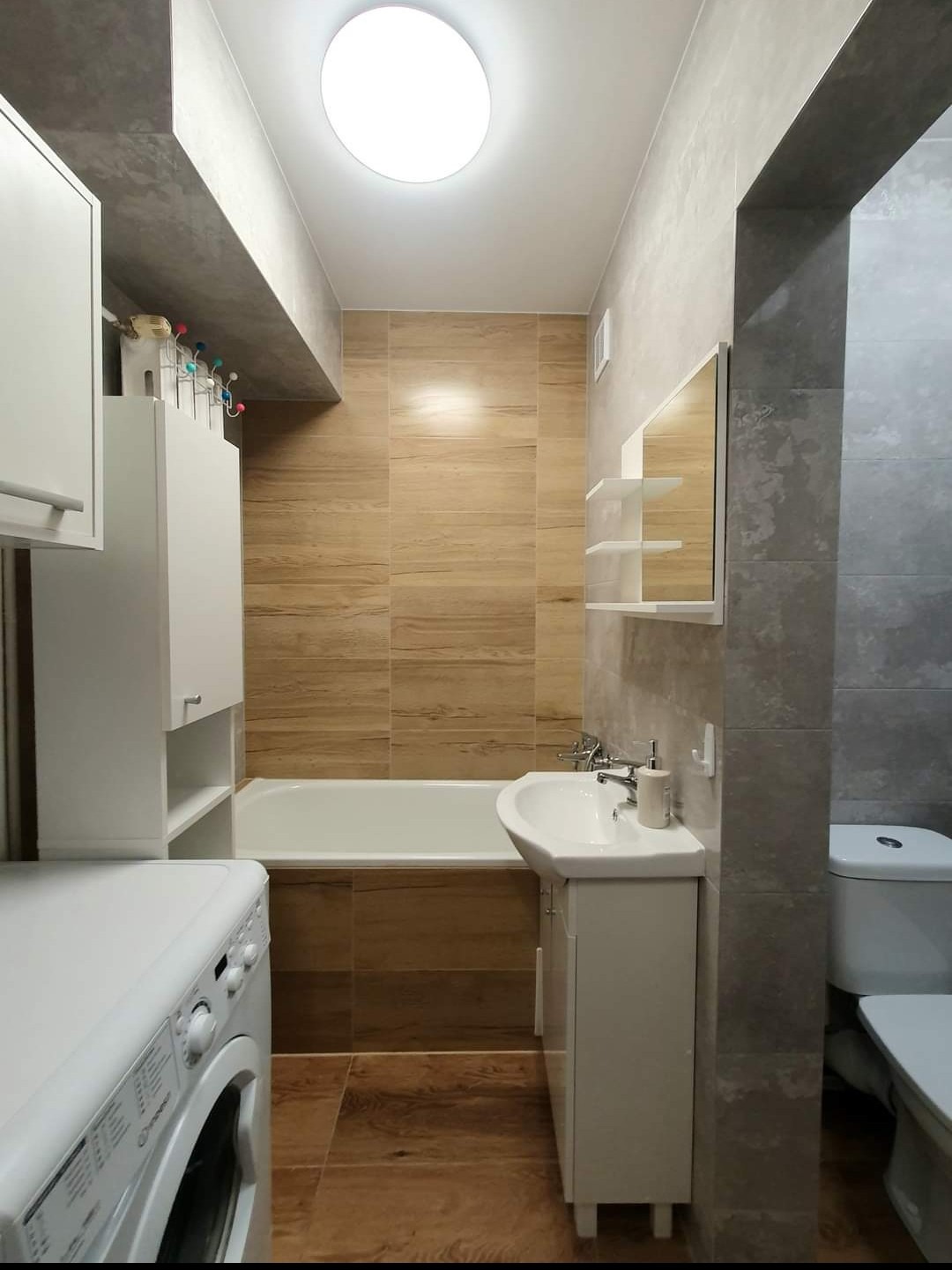 Ukończona łazienka z brązowymi płytkami imitującymi drewno, białą wanną, umywalką z szafką, pralką i toaletą, oświetlona okrągłym plafonem.