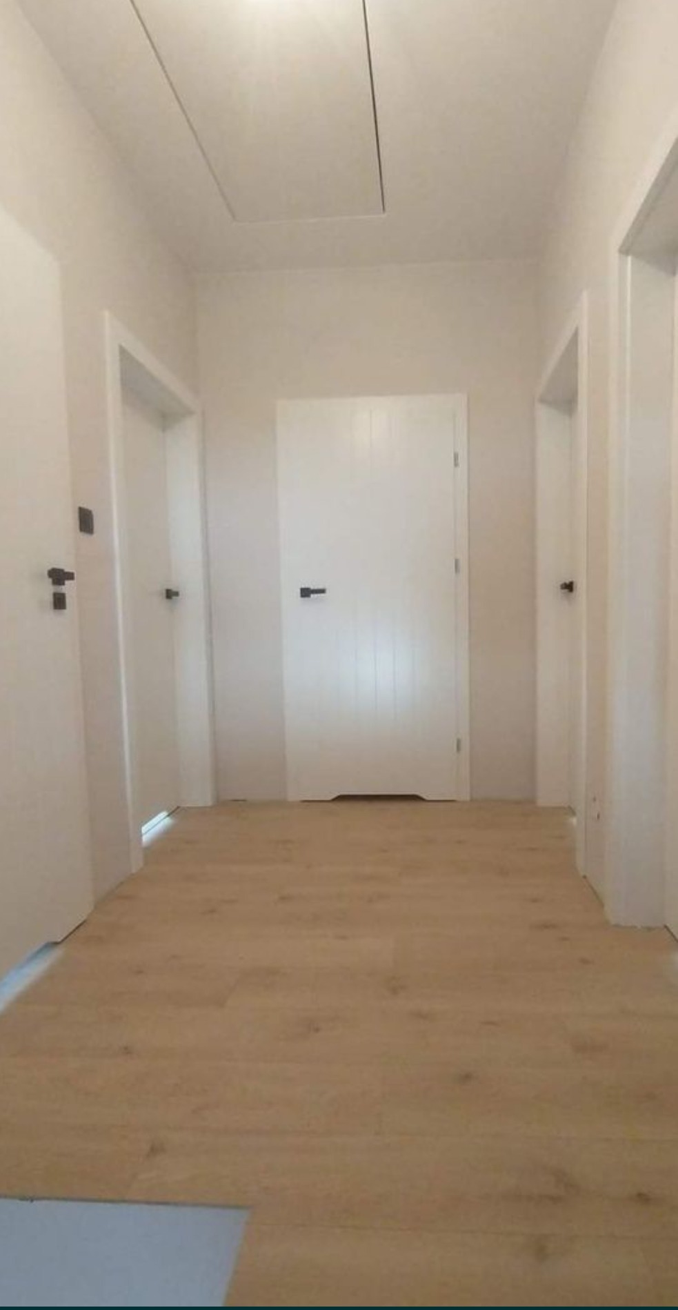 Wnętrze korytarza po remoncie z nową, jasną podłogą imitującą drewno i białymi drzwiami z czarnymi klamkami, widoczny właz na suficie.
