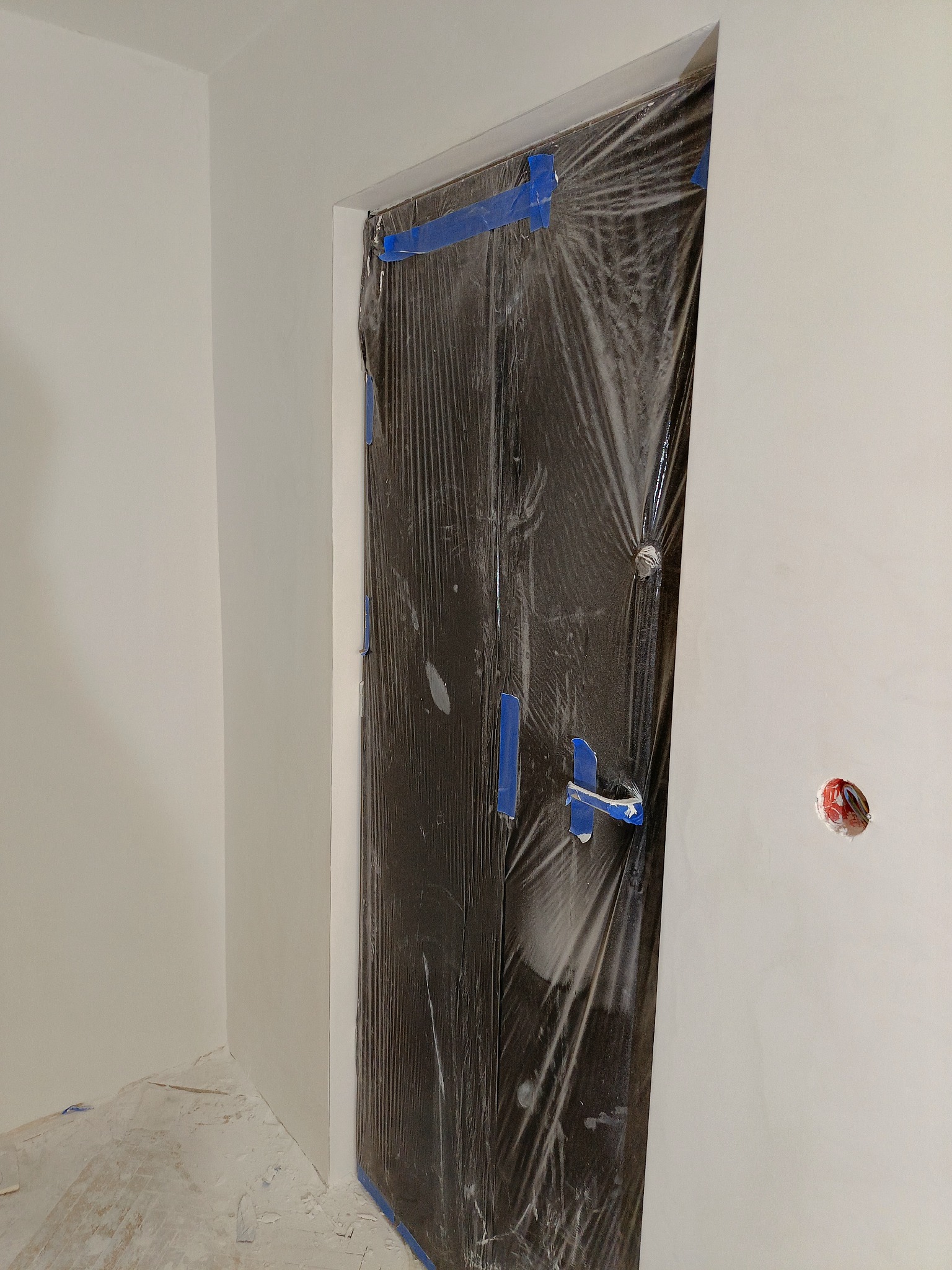 Drzwi zabezpieczone folią i niebieską taśmą malarską podczas prac malarskich na ścianach w jasnym odcieniu.