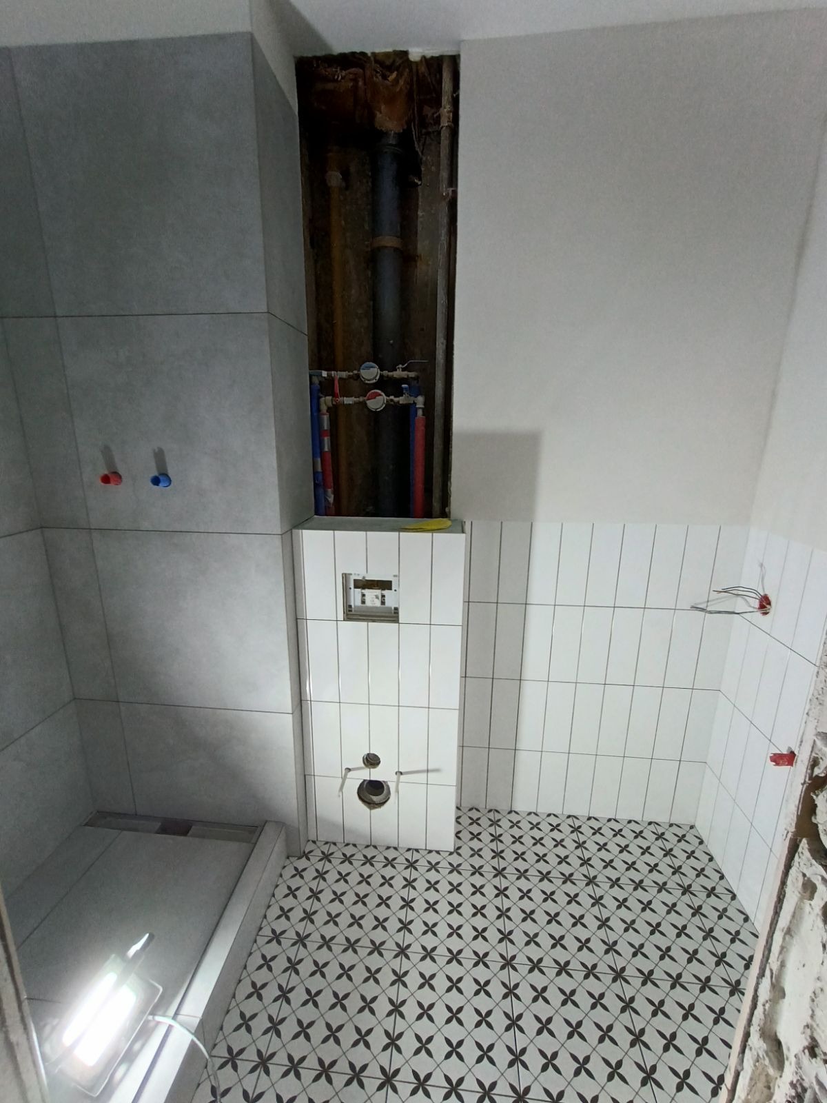 Wnętrze łazienki w trakcie prac: ułożone płytki podłogowe z motywem kwiatowym, częściowo wyłożone ściany białymi i szarymi płytkami, widoczne rury instalacji wodnej w ścianie, przygotowane punkty...