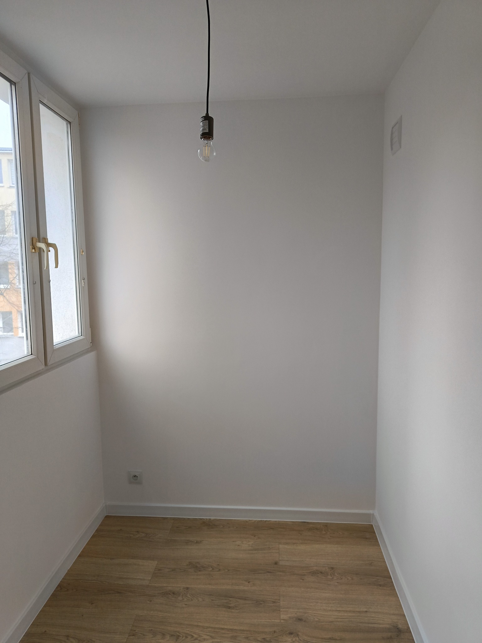 Puste, pomalowane na biało pomieszczenie w Łodzi z panelami podłogowymi, białymi listwami przypodłogowymi, oknem z widokiem na budynek i wiszącą żarówką na kablu.