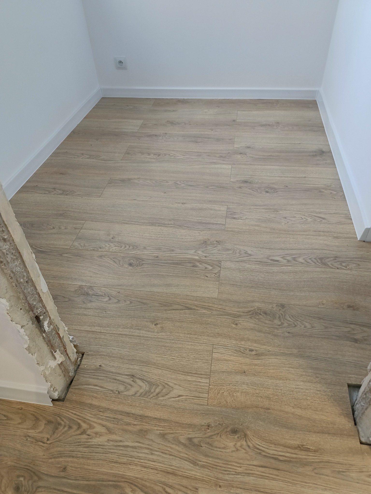 Jasne panele podłogowe imitujące drewno ułożone w pustym pokoju z białymi ścianami i listwami przypodłogowymi, widoczne surowe wykończenie ościeżnicy drzwi.