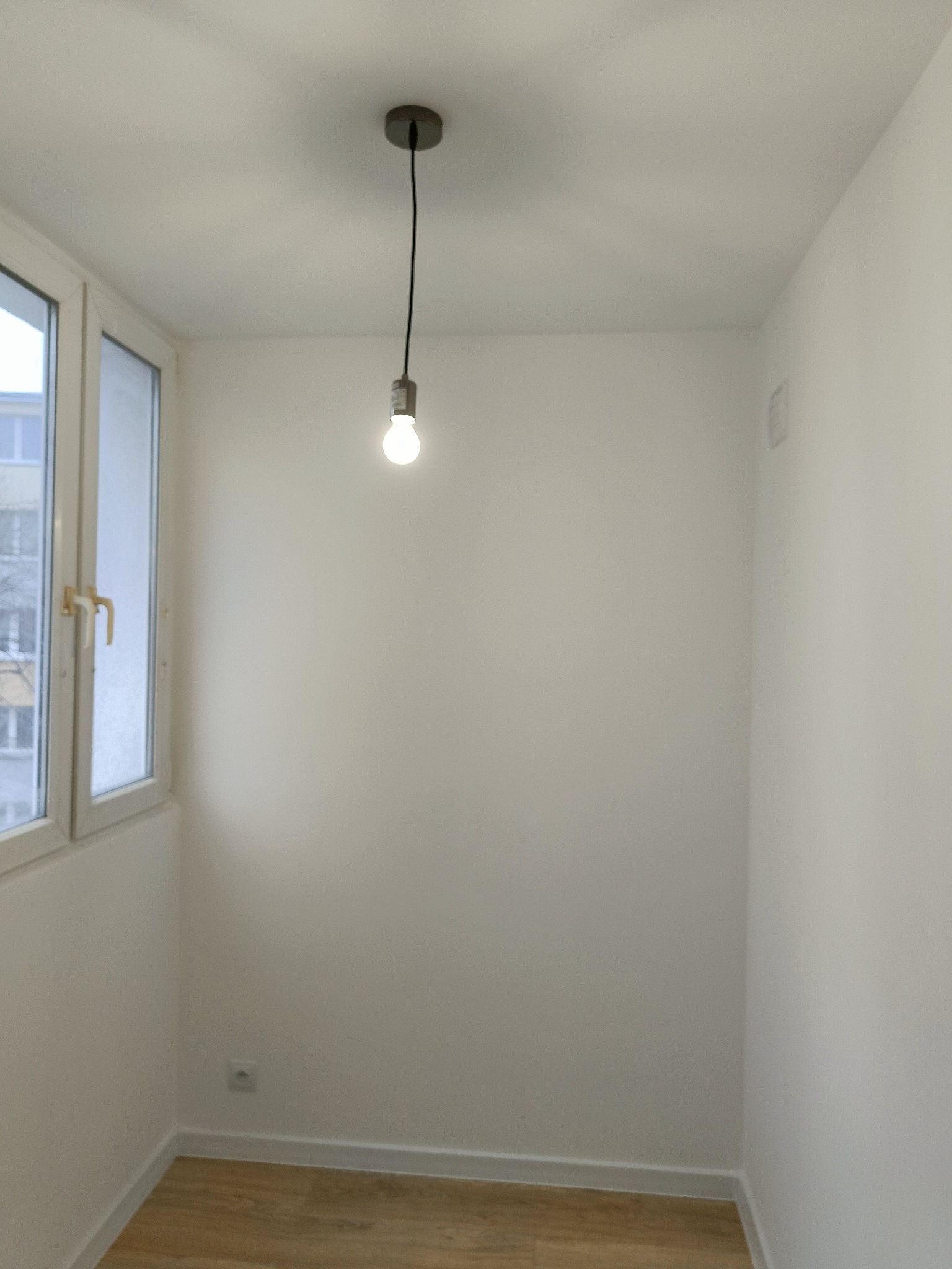 Pusty, odnowiony pokój z drewnianą podłogą, świeżo pomalowanymi białymi ścianami, oknem z widokiem na budynek i pojedynczą żarówką wiszącą na czarnym kablu.