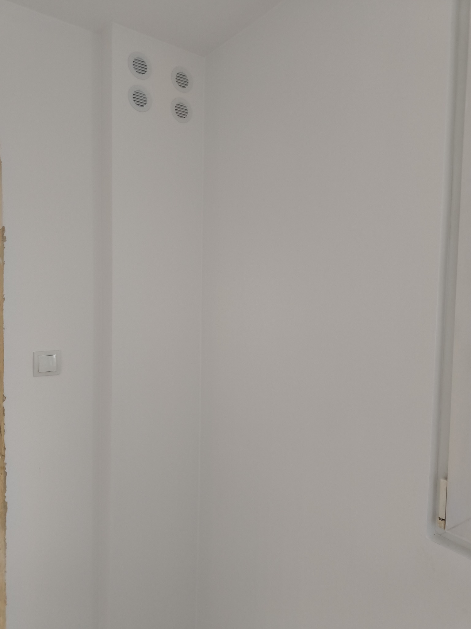 Świeżo pomalowana na biało ściana z czterema okrągłymi kratkami wentylacyjnymi w rogu i włącznikiem światła po lewej stronie, widoczny fragment ościeżnicy drzwi.