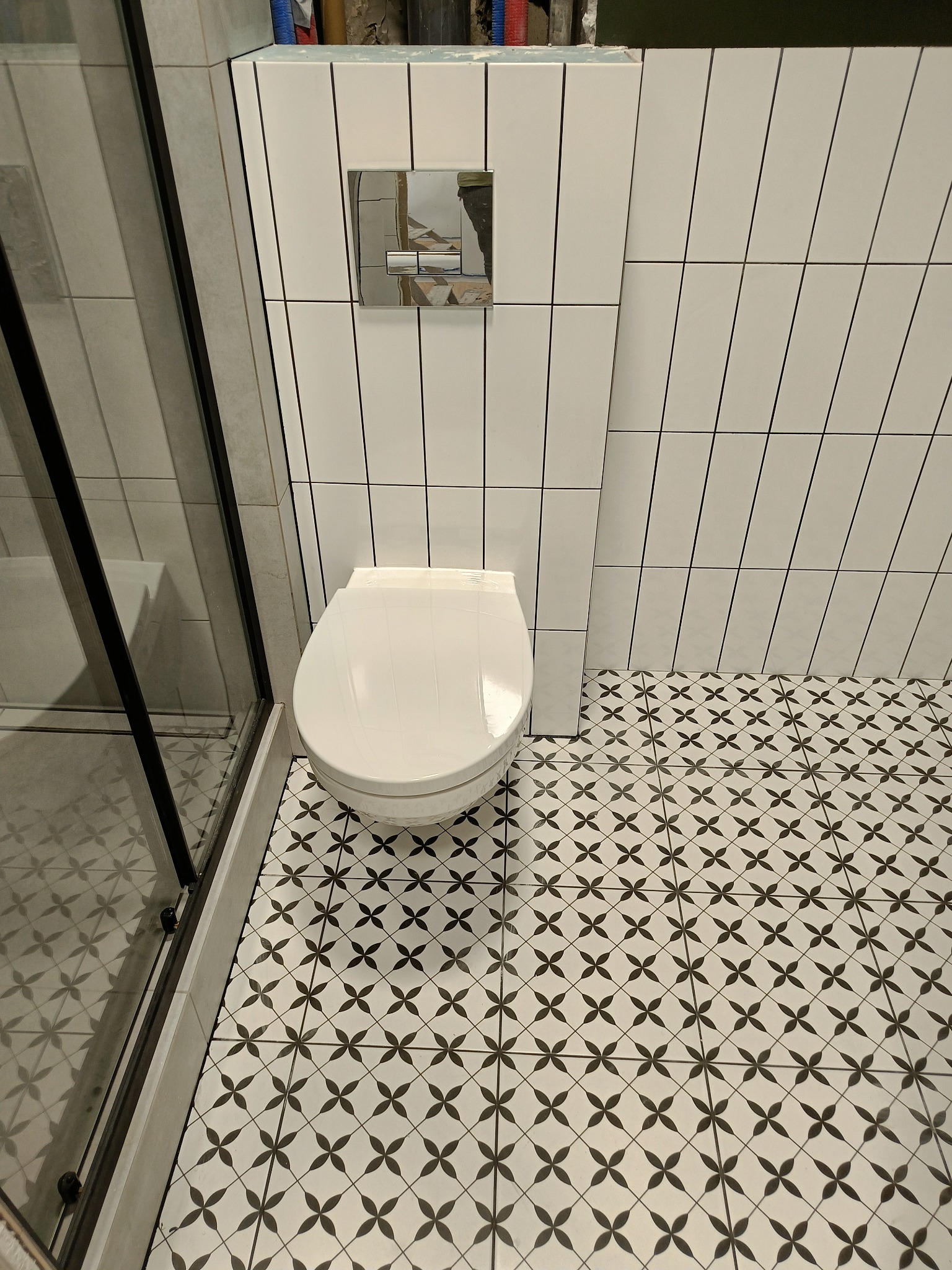 Nowoczesna łazienka z białą wiszącą toaletą, chromowanym przyciskiem spłukiwania na ścianie wyłożonej pionowymi białymi płytkami z czarną fugą oraz podłogą w czarno-białą geometryczną mozaikę...