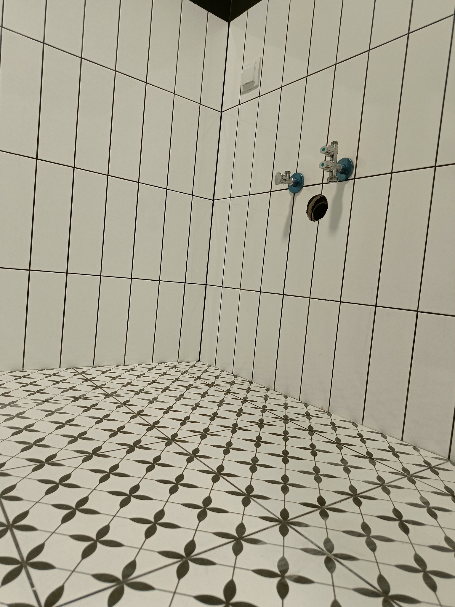 Wnętrze łazienki w trakcie prac wykończeniowych: białe, pionowo ułożone płytki ścienne typu 'metro', wzorzysta podłoga w czarno-białe kwiaty, przygotowane przyłącza wodne i elektryczne.