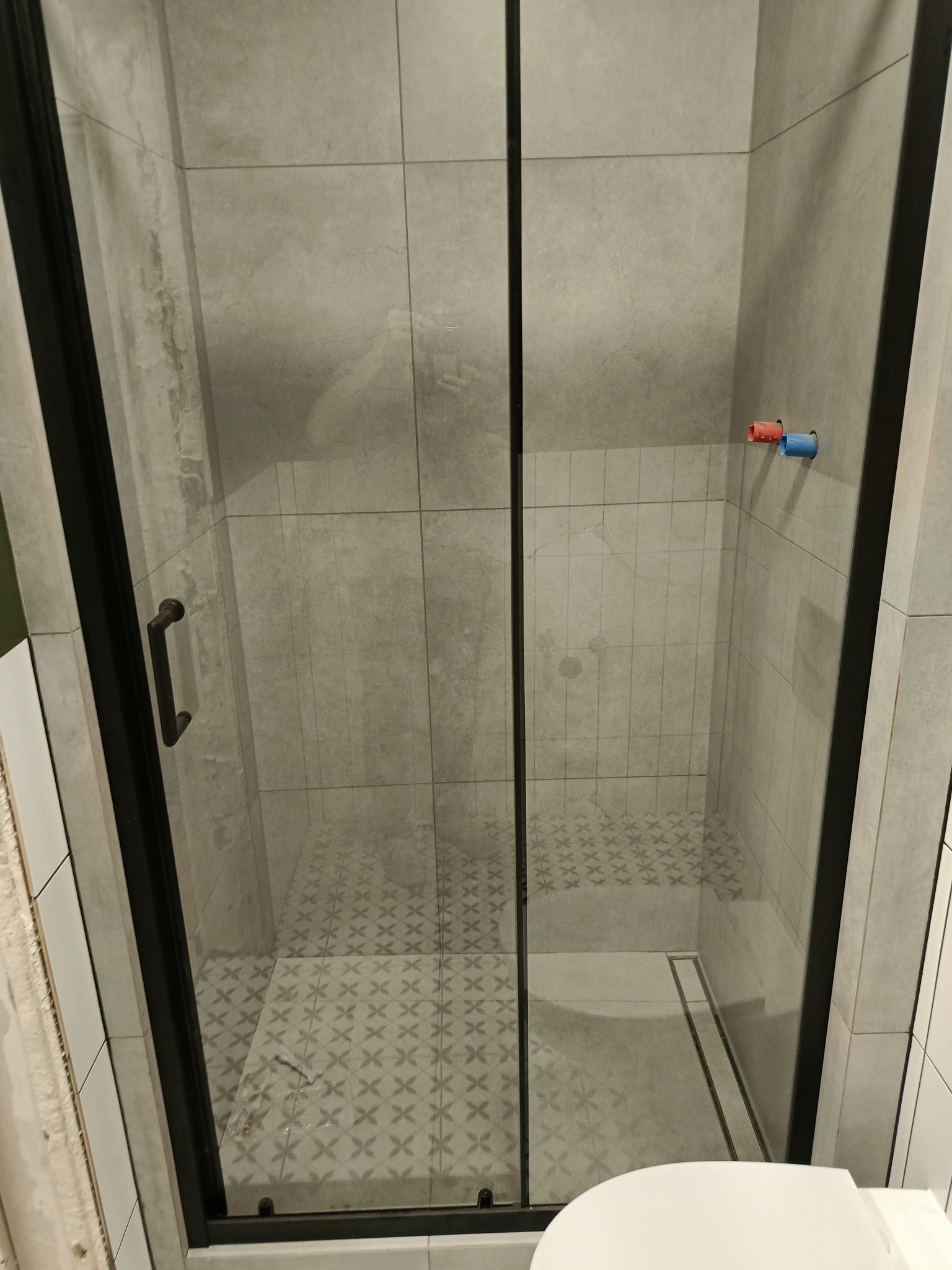 Nowoczesna kabina prysznicowa z czarną ramą i szklanymi drzwiami przesuwnymi, widoczne szare płytki na ścianach i podłodze z geometrycznym wzorem, z odpływem liniowym i fragmentem toalety w dolnej...