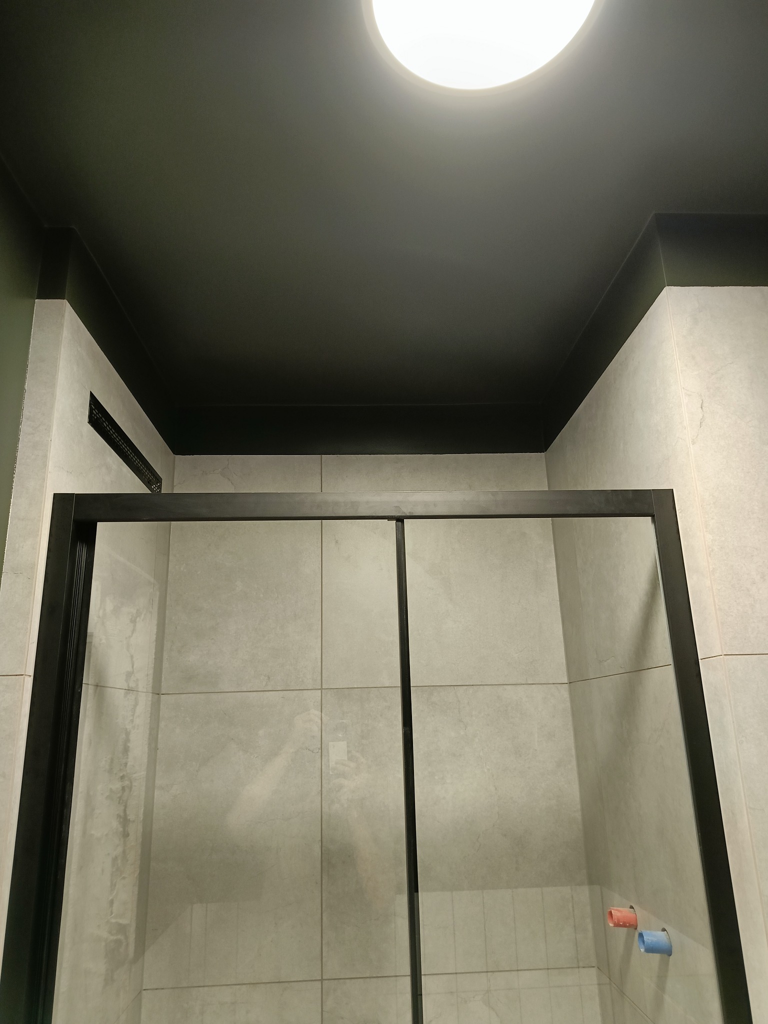 Nowoczesna kabina prysznicowa z czarną ramą, szare płytki na ścianach, ciemny sufit z okrągłym oświetleniem i kratką wentylacyjną, widoczne wystające rury z zaślepkami.