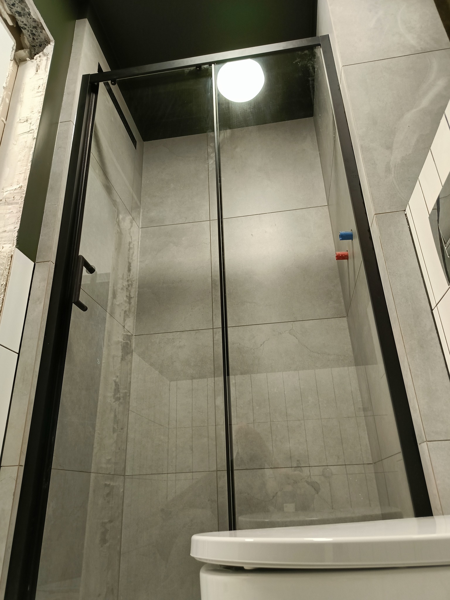 Nowoczesna kabina prysznicowa z czarnymi ramami i szarymi płytkami w trakcie montażu, widoczne niedokończone wykończenie ściany.