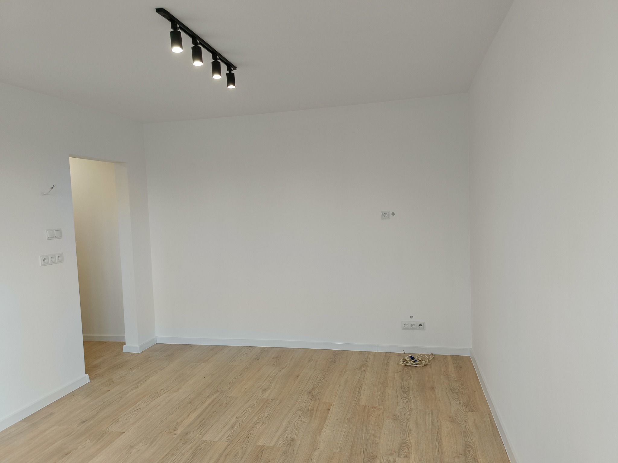 Puste, białe wnętrze z panelami podłogowymi w odcieniach drewna i czarną listwą oświetleniową na suficie. Na podłodze zwój kabli elektrycznych.