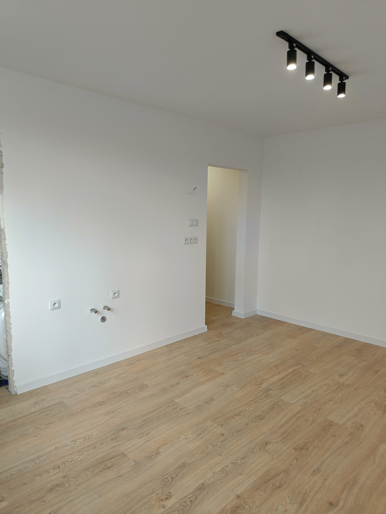Pusta, jasna przestrzeń w trakcie prac wykończeniowych: świeżo położona podłoga z paneli, białe ściany z widocznymi puszkami elektrycznymi i instalacją wodną, czarna listwa sufitowa z reflektorami.