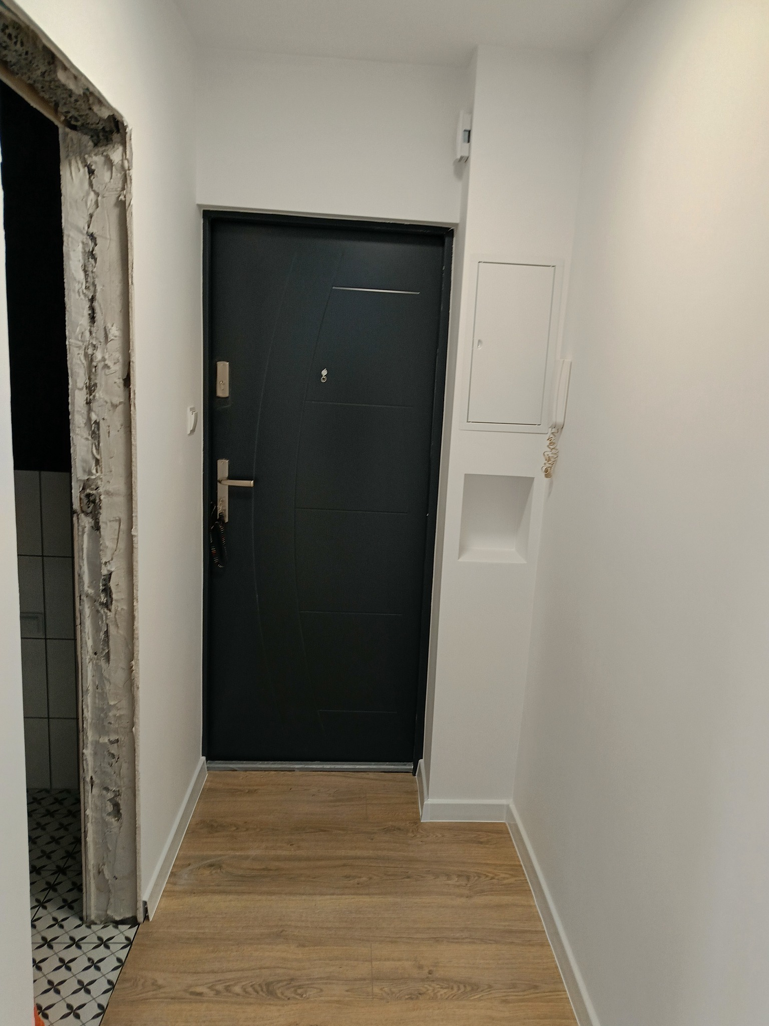 Korytarz po remoncie z ciemnymi drzwiami wejściowymi, jasnymi ścianami i panelami podłogowymi imitującymi drewno. Ościeżnica po lewej stronie w trakcie prac wykończeniowych, widoczna surowa ściana.