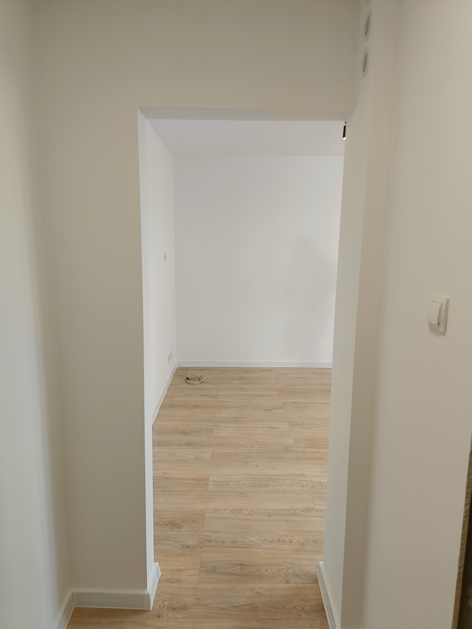 Puste, odświeżone wnętrze mieszkania z parkietem ułożonym w jodełkę, widoczne przez otwarte drzwi; na ścianie włącznik światła, na podłodze zwój kabli.