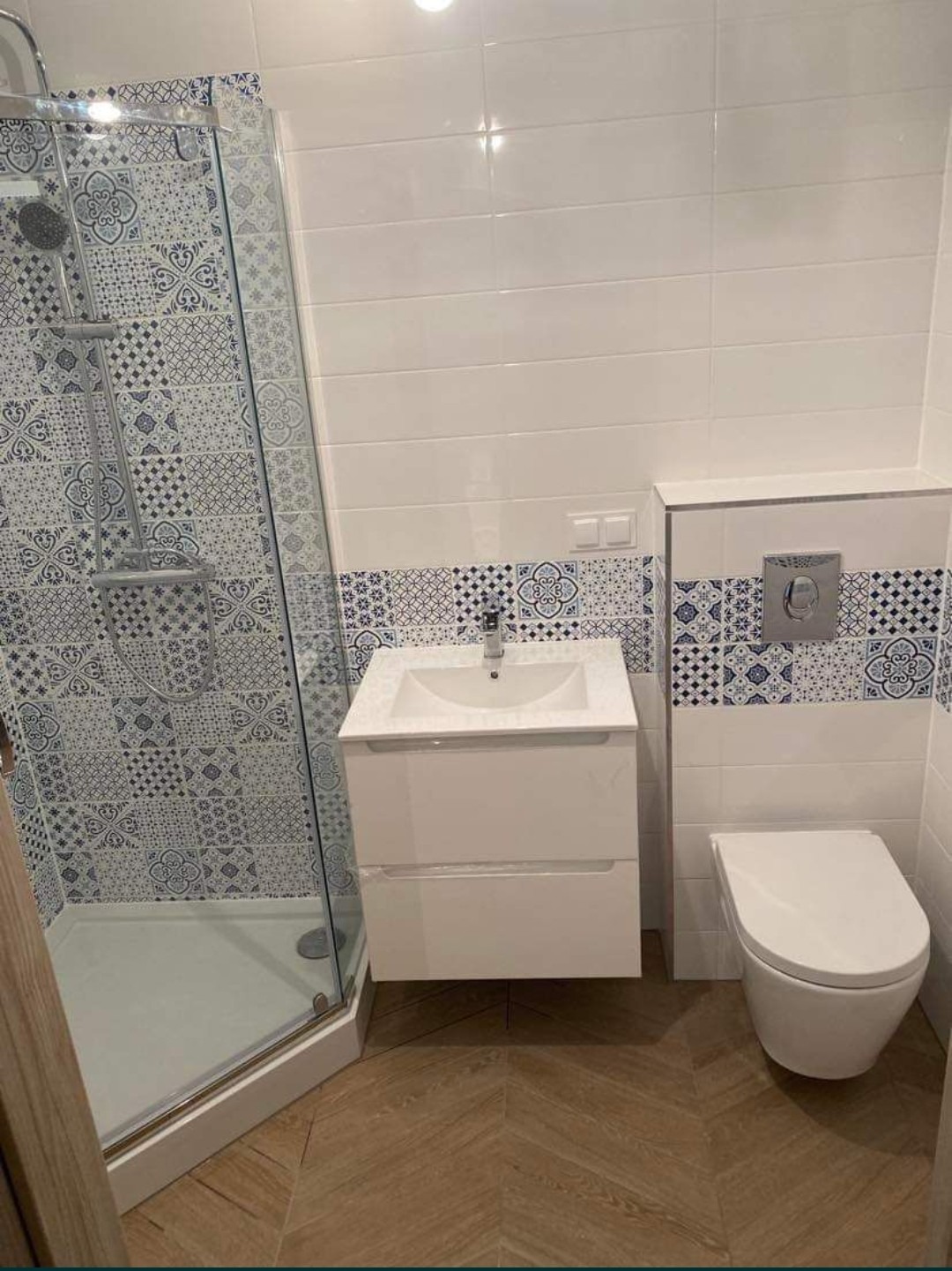 Nowoczesna łazienka z kabiną prysznicową z wzorzystymi płytkami, białą umywalką z szafką i podwieszaną toaletą, wykończona płytkami w stylu marokańskim i drewnopodobną podłogą ułożoną w jodełkę.