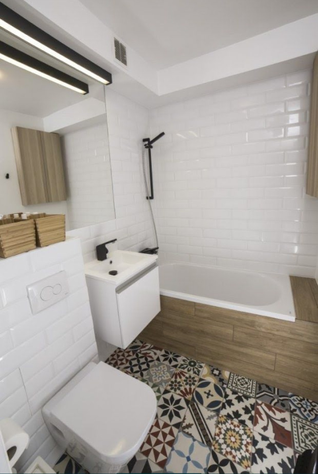 Nowoczesna łazienka z białą armaturą, wanną obudowaną drewnopodobnymi płytkami i wzorzystą, kolorową podłogą z płytek cementowych.