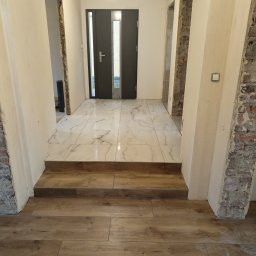 Usługi Ogólnobudowlane Paweł Pikus - Korytarz w trakcie remontu: położona podłoga z płytek imitujących drewno, białe płytki marmurowe na podwyższeniu, surowe ściany z fragmentami odkrytej cegły wokół otworów drzwiowych.