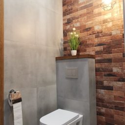Usługi Ogólnobudowlane Paweł Pikus - Nowoczesna łazienka z wiszącą toaletą, szarą płytką imitującą beton i ścianą z cegiełek, oświetlona designerską lampą z metalowym kloszem.