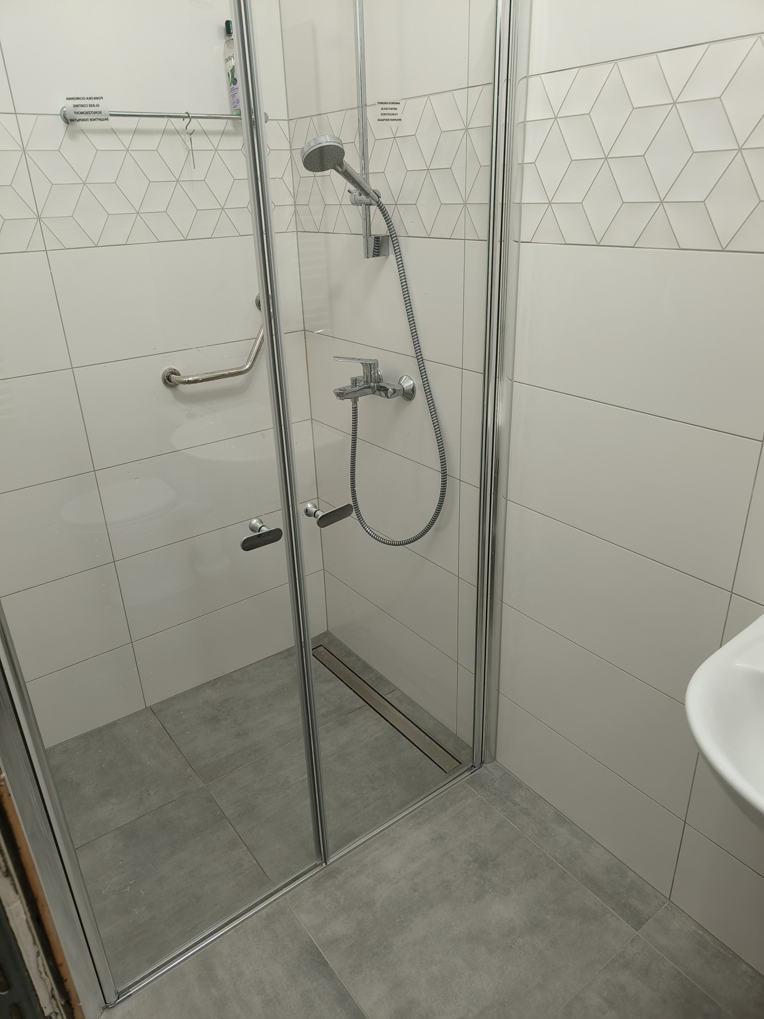 Wykończona łazienka z kabiną prysznicową ze srebrnymi okuciami, szarymi płytkami podłogowymi i białymi płytkami ściennymi z geometrycznym wzorem, uchwyt dla osób z niepełnosprawnościami zamontowany...