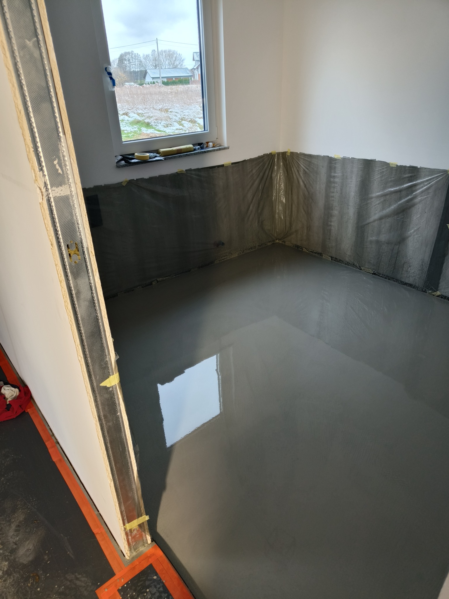Świeżo wylana, gładka wylewka samopoziomująca w pomieszczeniu zabezpieczonym folią malarską przy ścianach, widoczne profile przyścienne i okno z widokiem na zaśnieżony krajobraz.