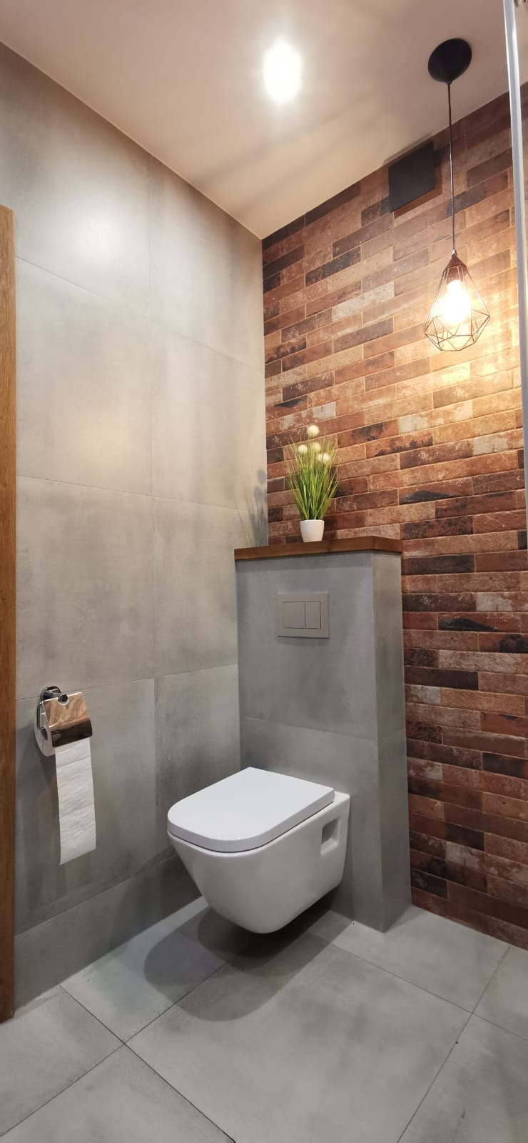 Nowoczesna łazienka z wiszącą toaletą, szarą płytką imitującą beton i ścianą z cegiełek, oświetlona designerską lampą z metalowym kloszem.
