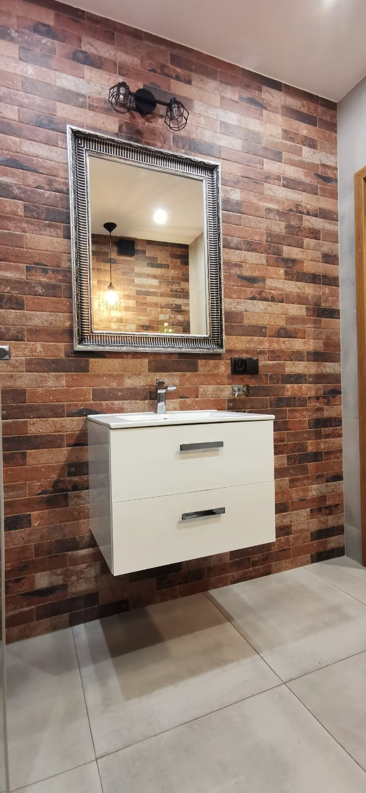 Nowoczesna łazienka z umywalką nablatową na szafce z dwiema szufladami, lustrem w ozdobnej ramie i ścianą wyłożoną cegiełkami w odcieniach brązu i czerni.