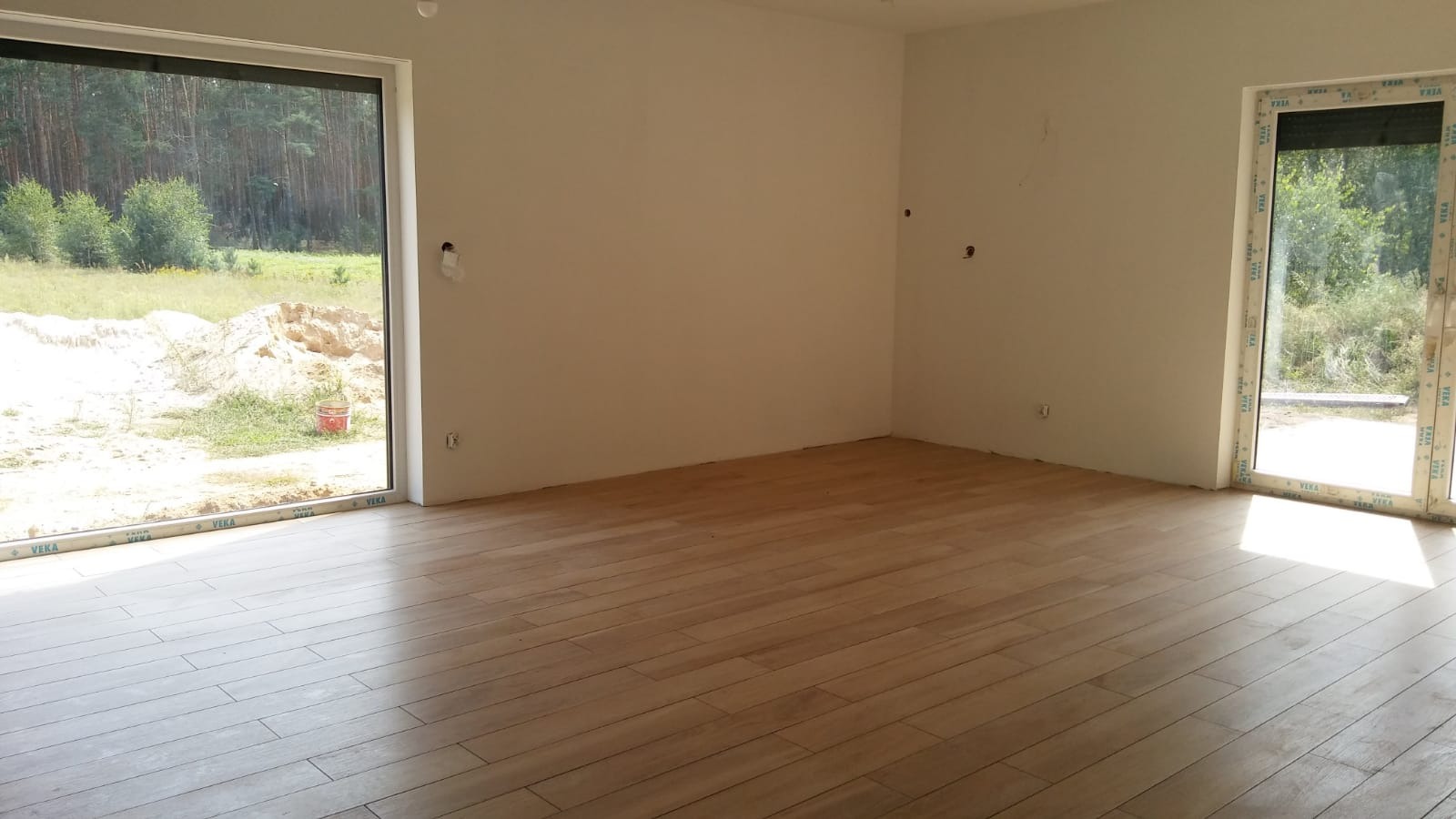 Jasne panele podłogowe ułożone w nowym pomieszczeniu z dużymi oknami, widoczne ślady po pracach budowlanych na ścianach.