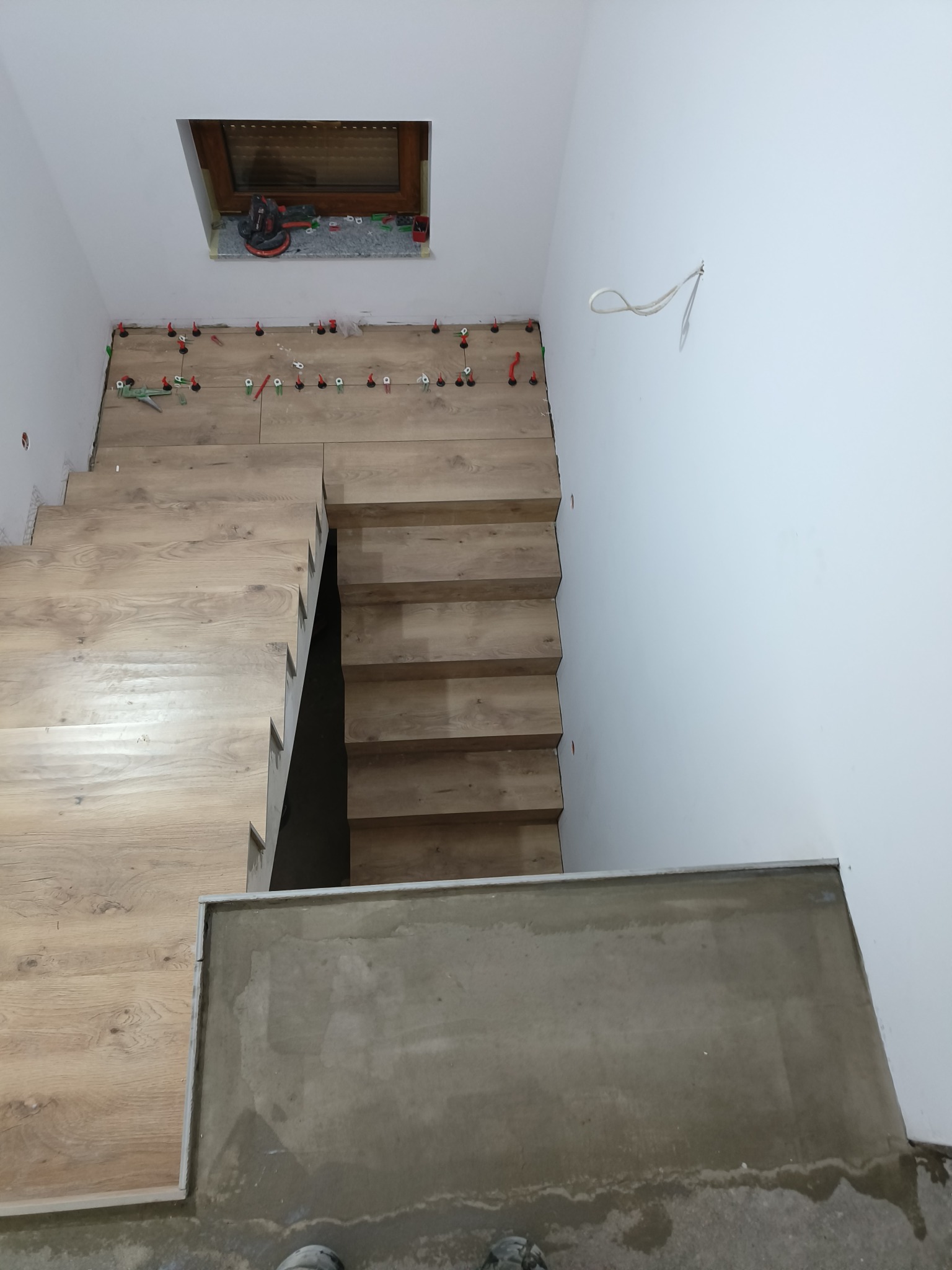 Montaż drewnopodobnych paneli na schodach, widoczne narzędzia i system poziomowania, surowe ściany w tle.