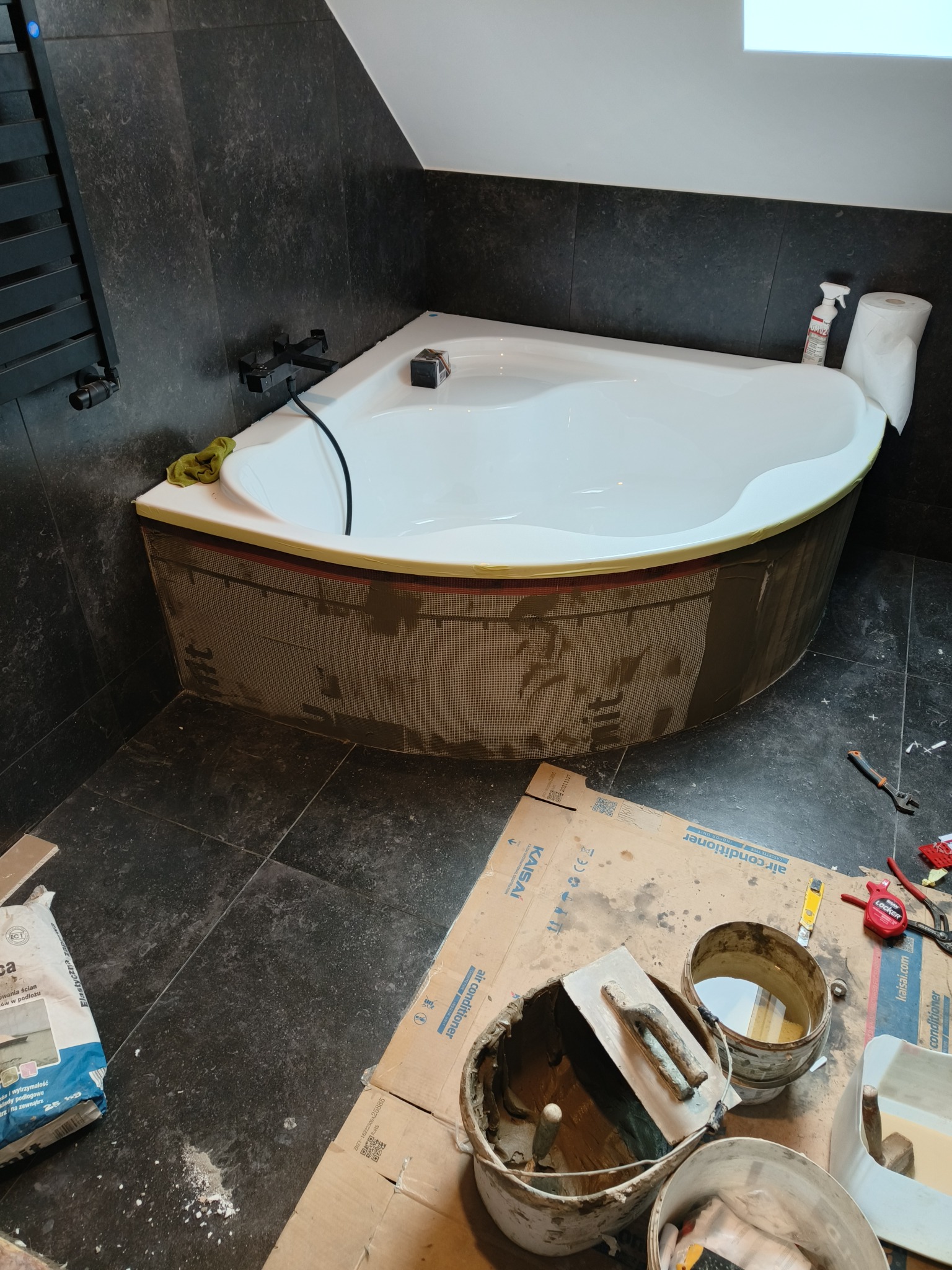 Narożna wanna w trakcie obudowy mozaiką w łazience z ciemnymi płytkami; widoczne narzędzia glazurnicze, klej i kartonowe zabezpieczenie podłogi.