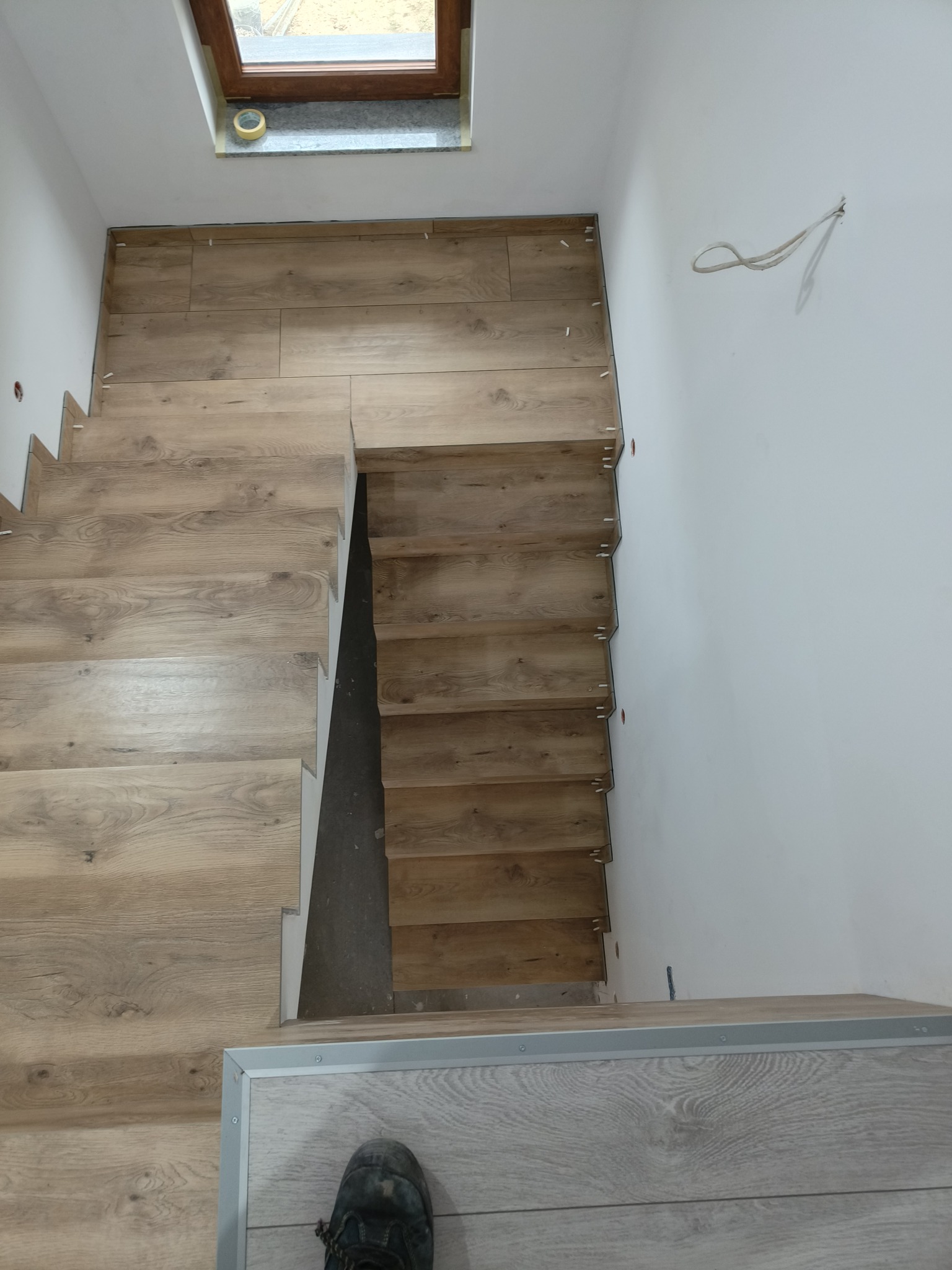 Montaż drewnopodobnych płytek na schodach wewnętrznych, widoczne plastikowe krzyżyki dystansowe i fragment roboczego buta w dolnym rogu.