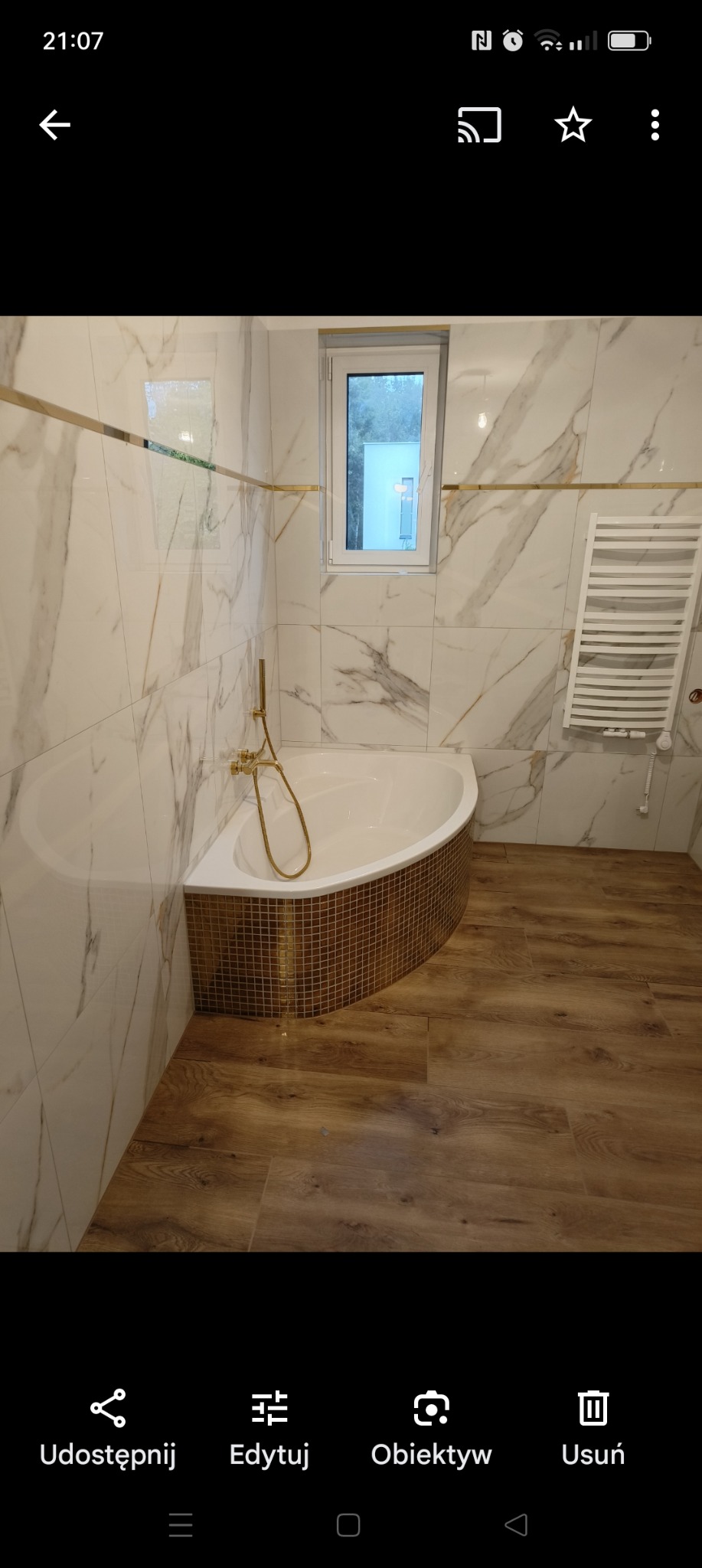 Nowoczesna łazienka z narożną wanną obudowaną mozaiką w odcieniach złota, marmurowe płytki na ścianach z dekoracyjną złotą listwą, drewnopodobna podłoga, biały grzejnik drabinkowy i okno z widokiem...