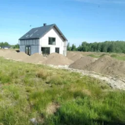 Wyr&oacute;wnany teren pod budowę domu jednorodzinnego, widok z kabiny maszyny, hałdy ziemi przed budynkiem w stanie surowym zamkniętym, niebo bezchmurne.