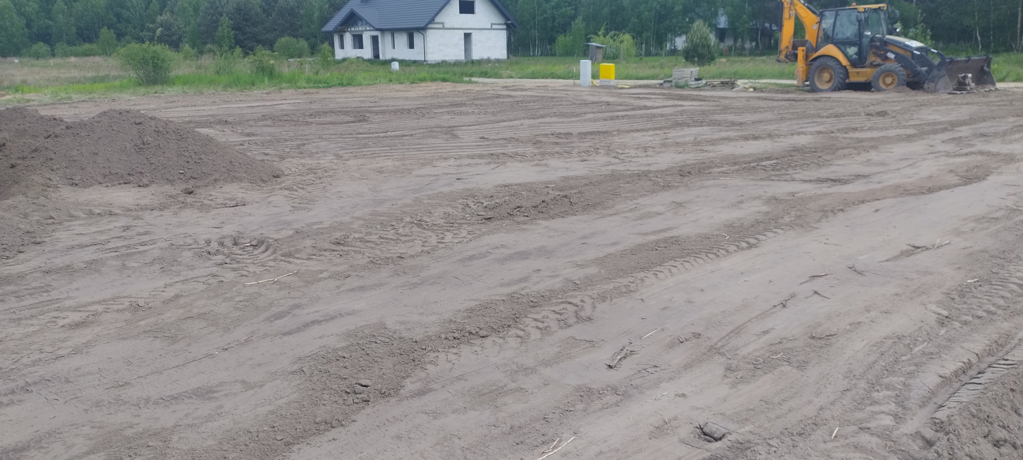 Przygotowany teren pod budowę domu jednorodzinnego z żółtą koparko-ładowarką w tle, widoczne ślady opon i pryzma ziemi.