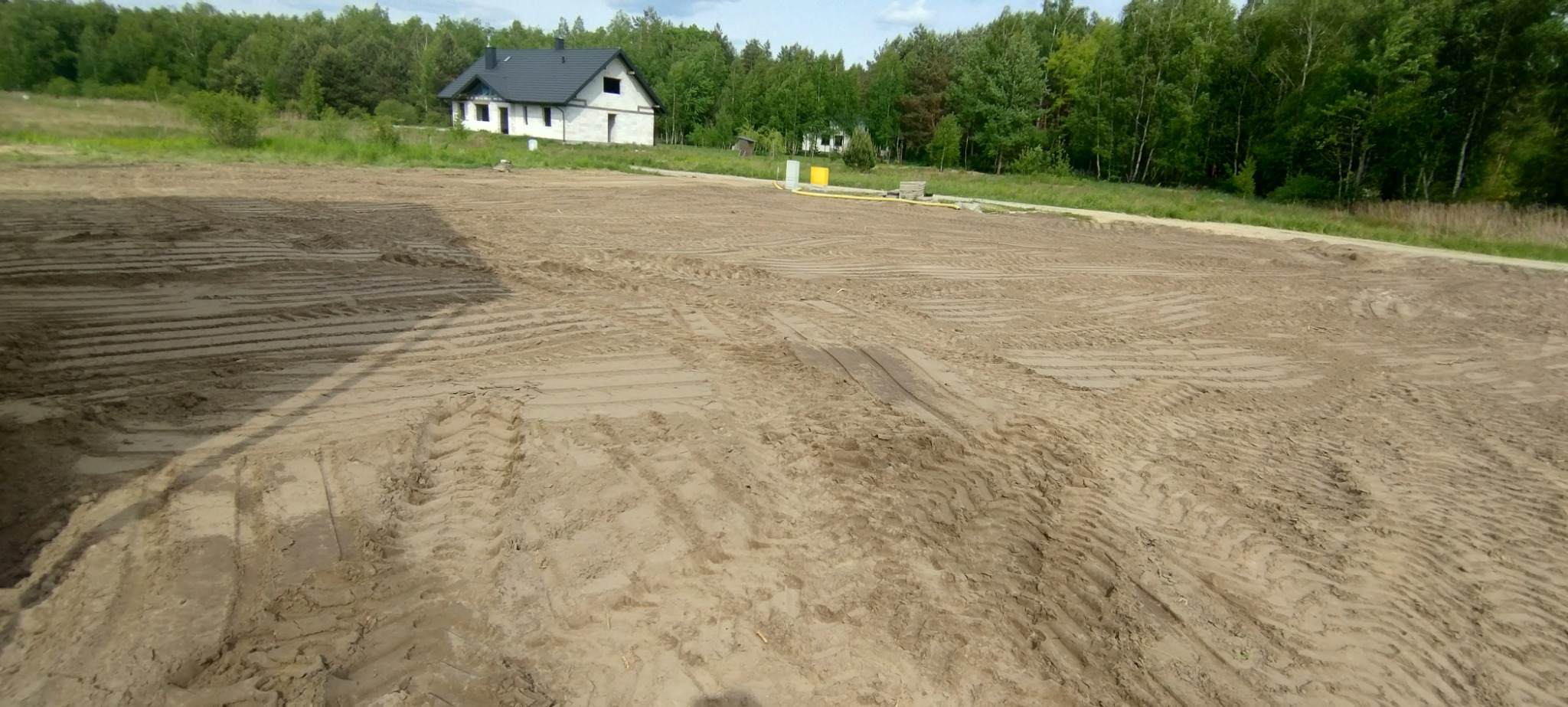 Przygotowany teren pod budowę domu jednorodzinnego, wyrównany piasek z wyraźnymi śladami opon ciężkiego sprzętu, w tle biały dom w stanie surowym otoczony zielenią.