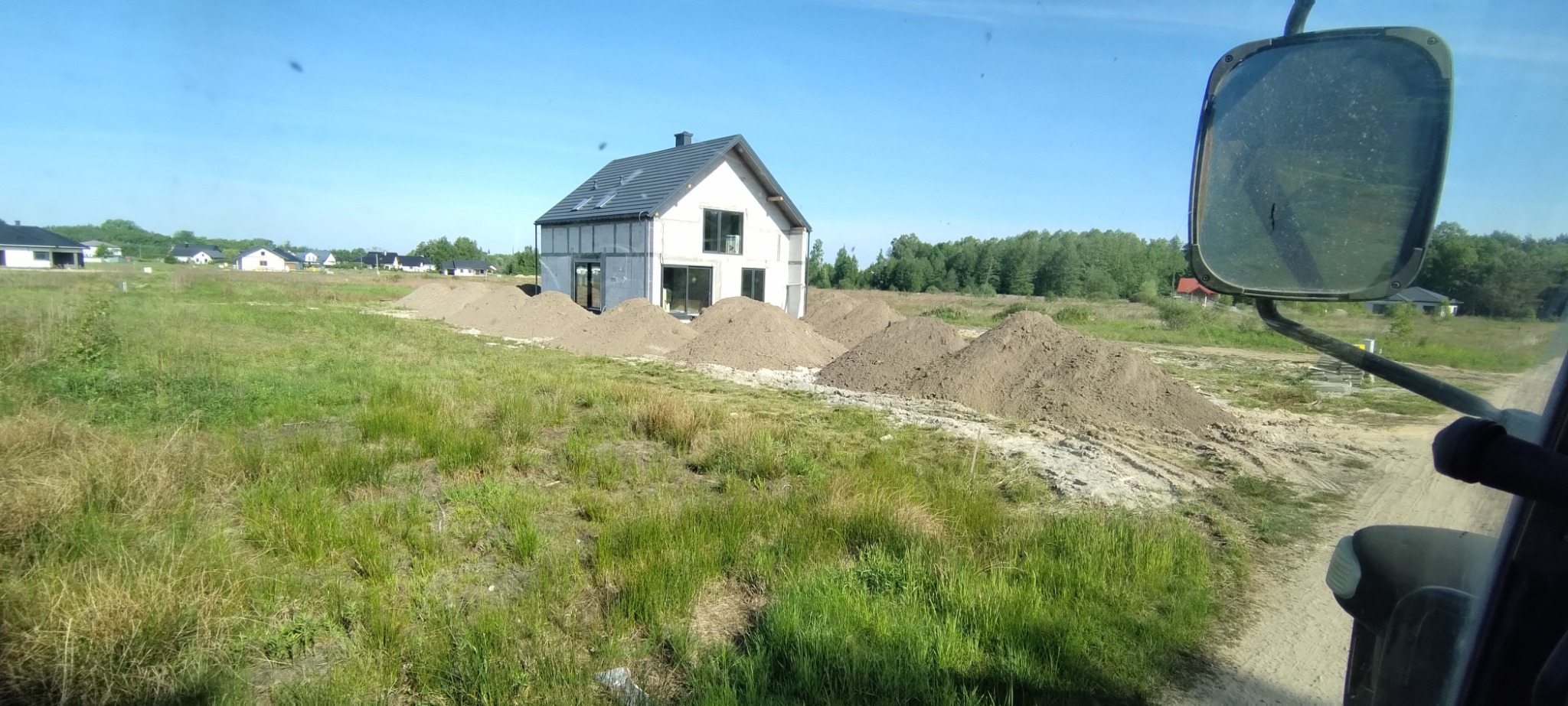 Wyrównany teren pod budowę domu jednorodzinnego, widok z kabiny maszyny, hałdy ziemi przed budynkiem w stanie surowym zamkniętym, niebo bezchmurne.