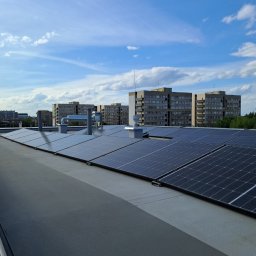 ELECTRICAL SOLUTIONS YK - Rząd paneli słonecznych na dachu budynku z szarym pokryciem, w tle widoczne bloki mieszkalne i błękitne niebo z chmurami.