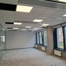 Wnętrze biurowe w stanie deweloperskim z widocznymi panelami sufitowymi z oświetleniem LED, instalacją przeciwpożarową, grzejnikami pod oknami i wykładziną dywanową, przygotowane do dalszego...