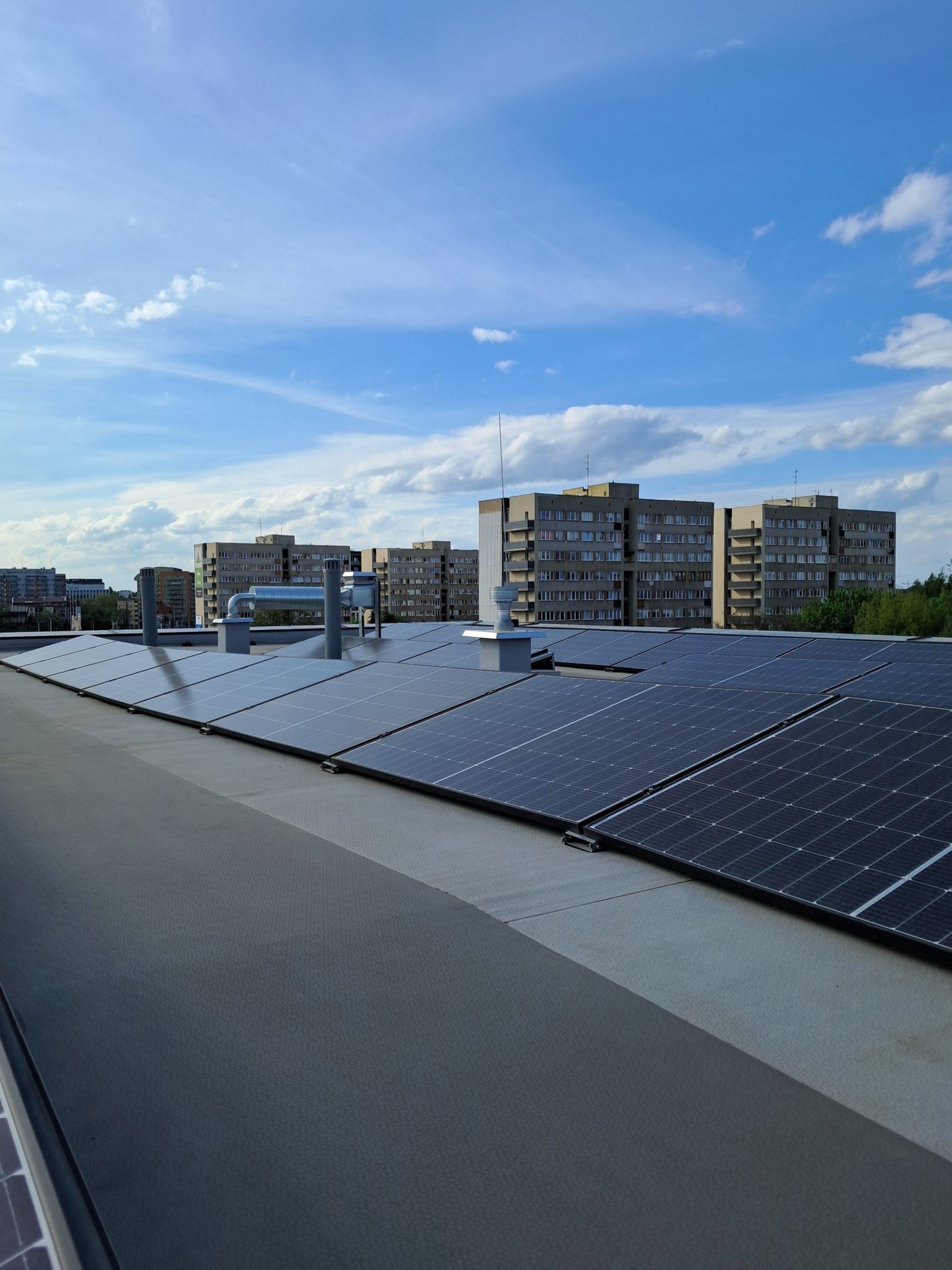 Rząd paneli słonecznych na dachu budynku z szarym pokryciem, w tle widoczne bloki mieszkalne i błękitne niebo z chmurami.