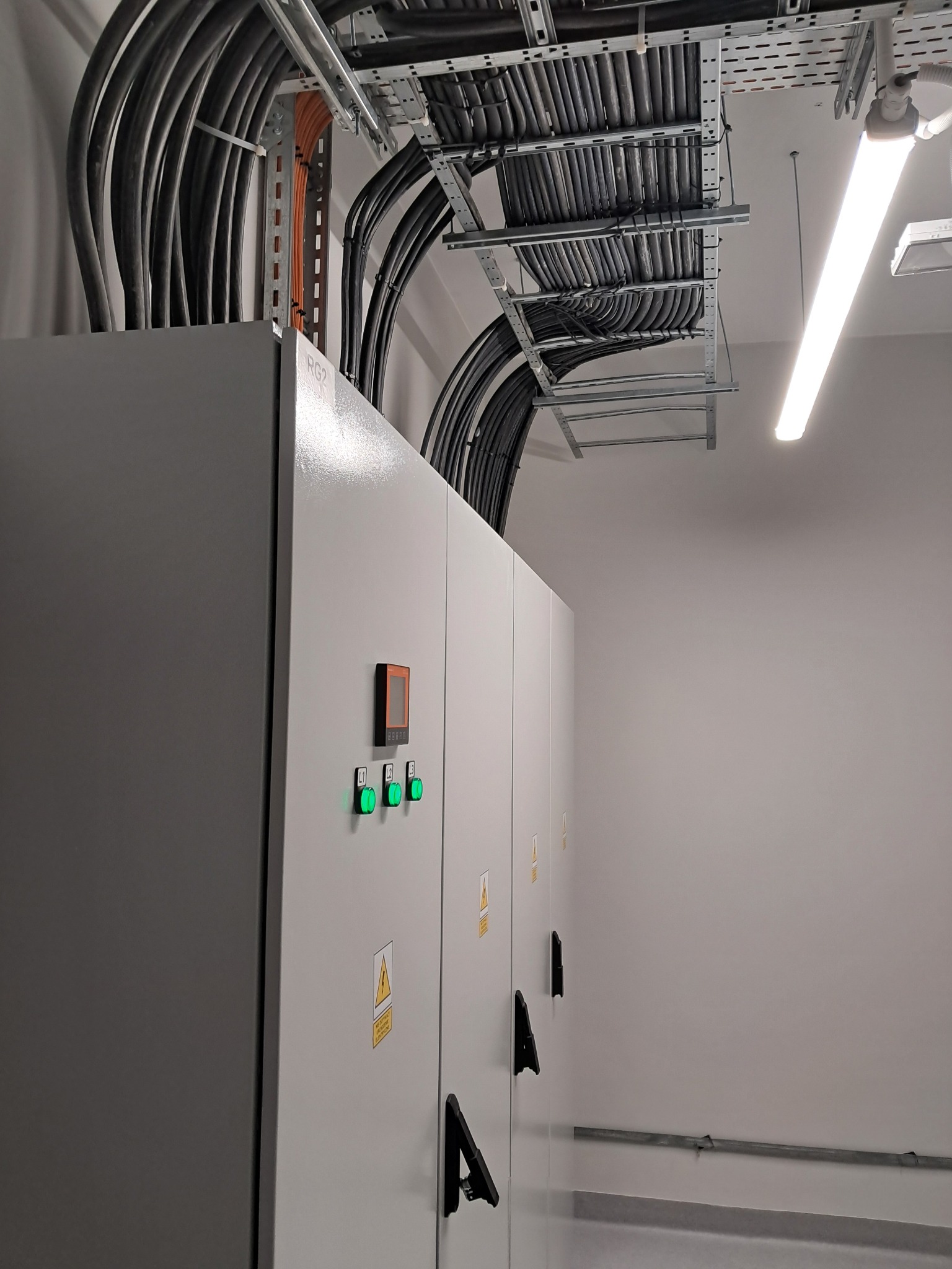 Szafa sterownicza z widocznymi przewodami elektrycznymi ułożonymi w korytach kablowych pod sufitem, z panelami kontrolnymi i ostrzeżeniami o napięciu.