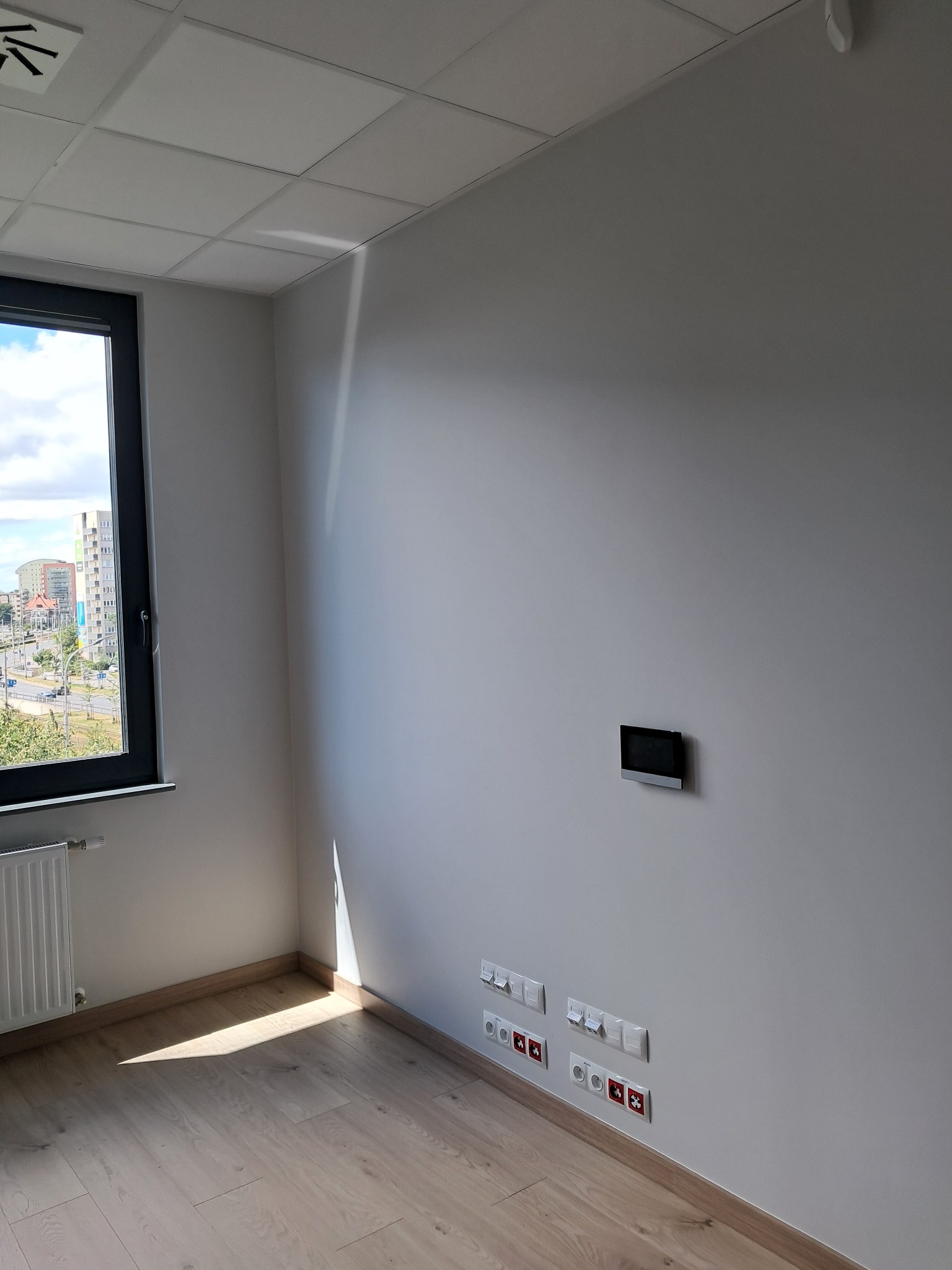 Wnętrze pokoju z widocznymi gniazdkami elektrycznymi, włącznikami światła oraz panelem sterowania alarmem na ścianie, parkiet na podłodze, okno z widokiem na miasto.