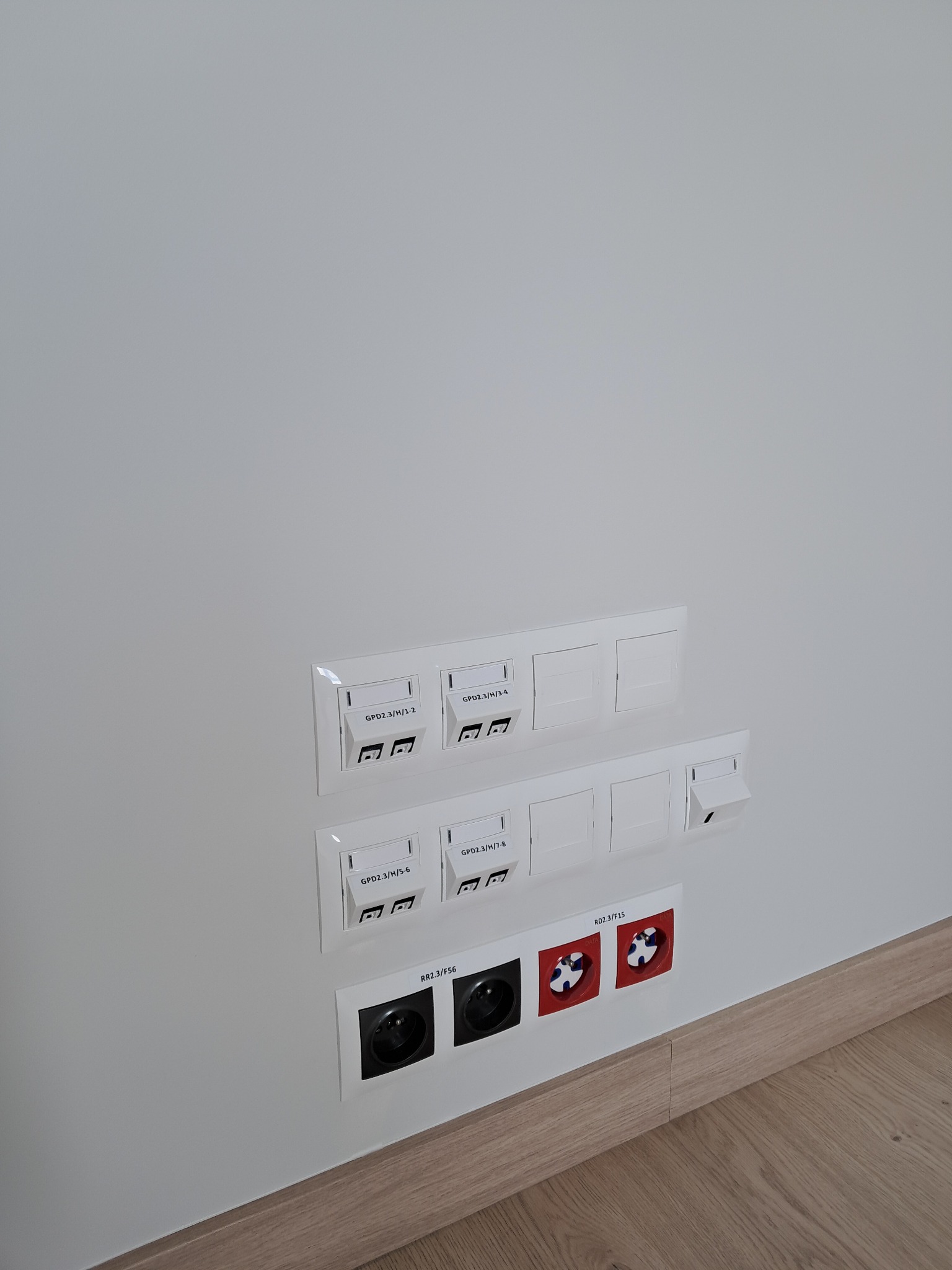 Biała ściana z zamontowanymi gniazdkami elektrycznymi, gniazdami sieciowymi i gniazdami alarmowymi, wszystkie w białych ramkach, z opisami nadrukowanymi na ramkach, zamontowane blisko podłogi...