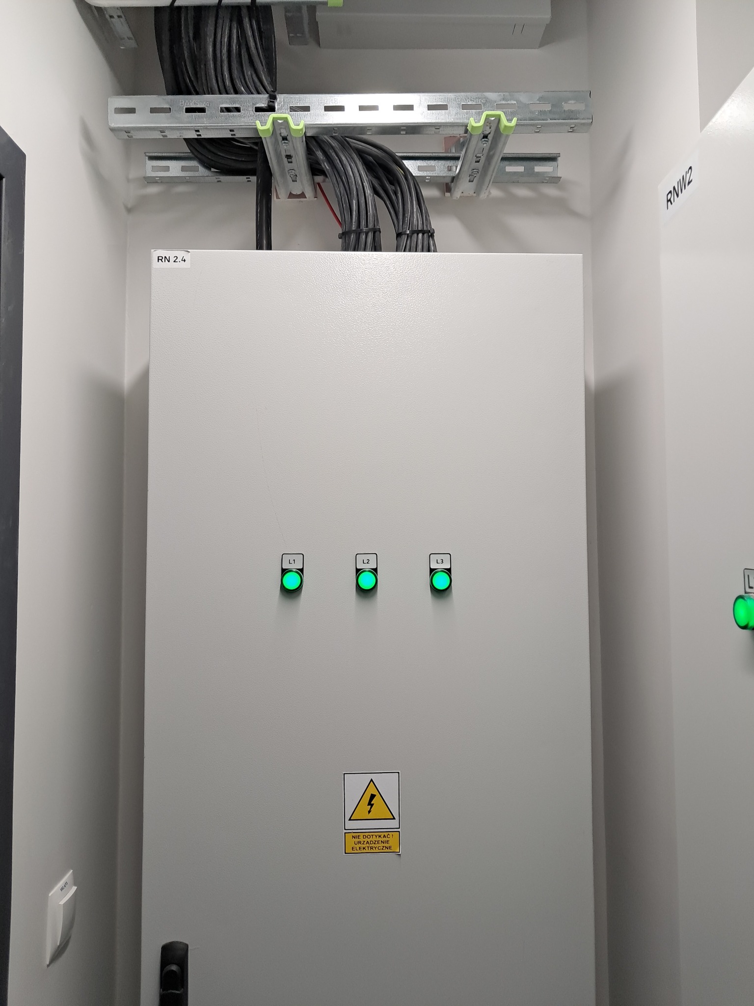 Szara szafa elektryczna z trzema świecącymi na zielono diodami (L1, L2, L3) i żółtą naklejką ostrzegawczą, widoczne kable w korytkach pod sufitem, oświetlenie górne, oznaczenie RN 2.4 na szafie...
