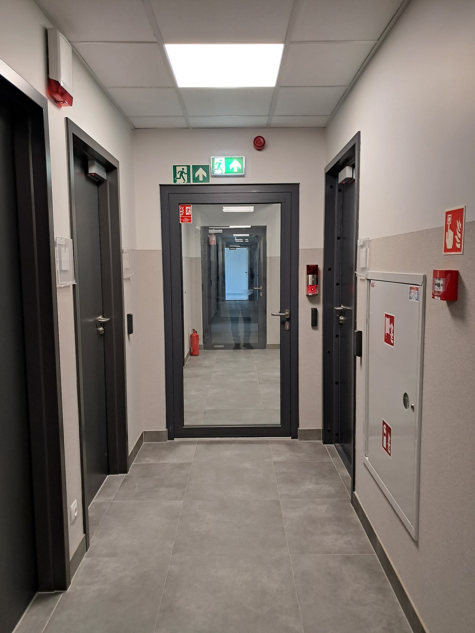 Korytarz w budynku z szarymi drzwiami, płytkami podłogowymi i systemem przeciwpożarowym, w tym gaśnicą, skrzynką na węża i alarmem.