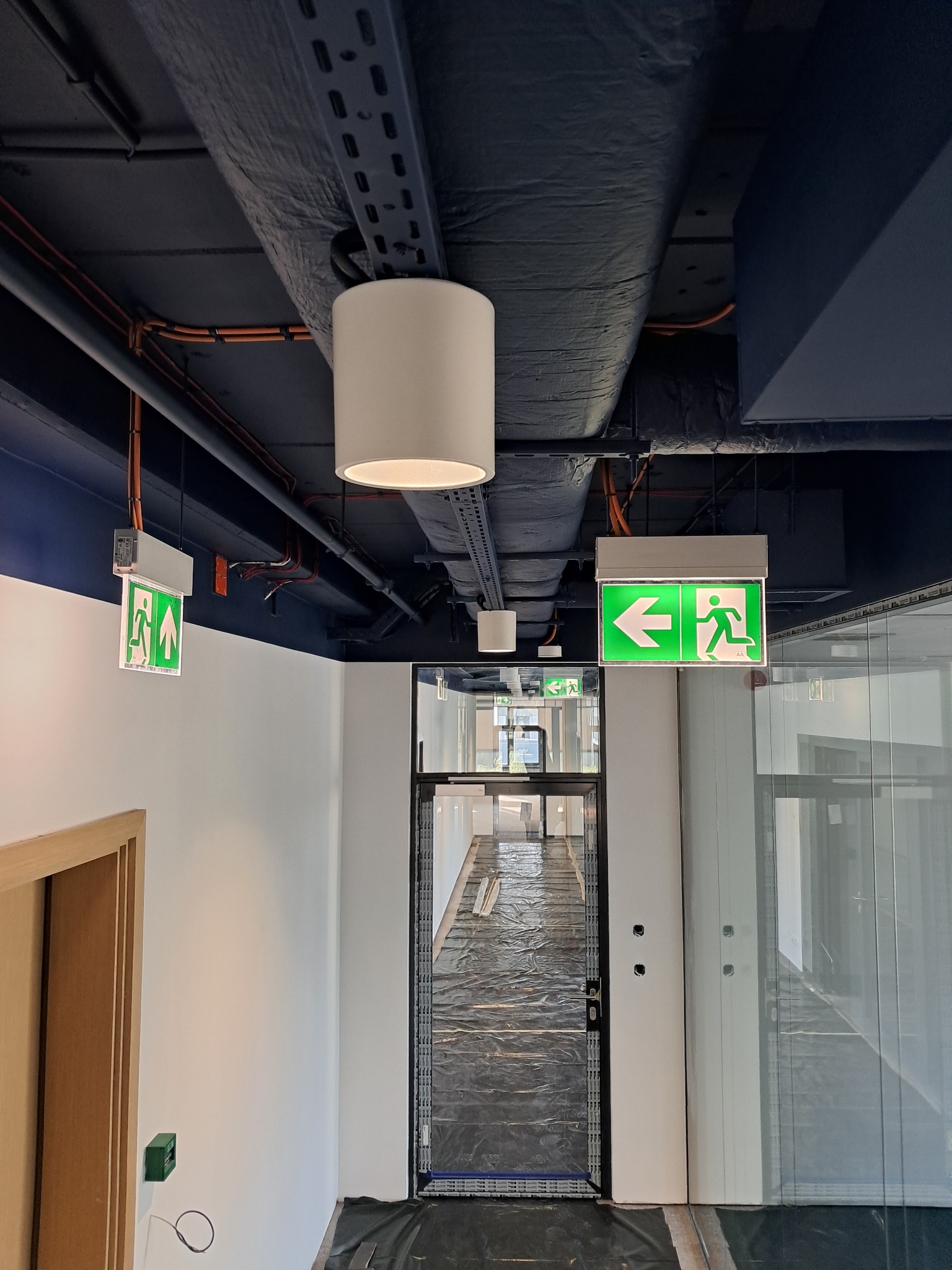 Korytarz biurowy w trakcie remontu z zamontowanym oświetleniem awaryjnym i widocznymi przewodami elektrycznymi pod sufitem.